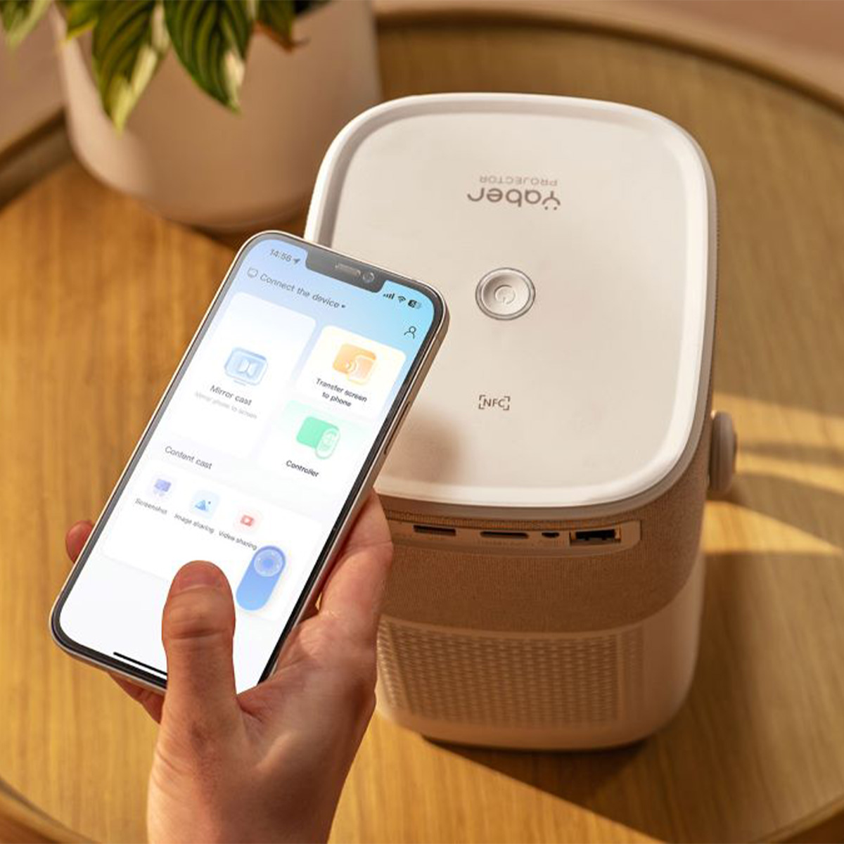 Osoba trzymająca telefon z aplikacją w pobliżu biało-beżowego projektora na stole.