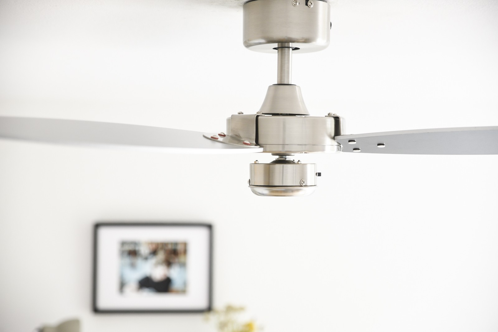 Nahaufnahme eines silbernen Deckenventilators. Die Blätter erstrecken sich zu den Seiten. Verschwommener Bilderrahmen und Wand im Hintergrund.