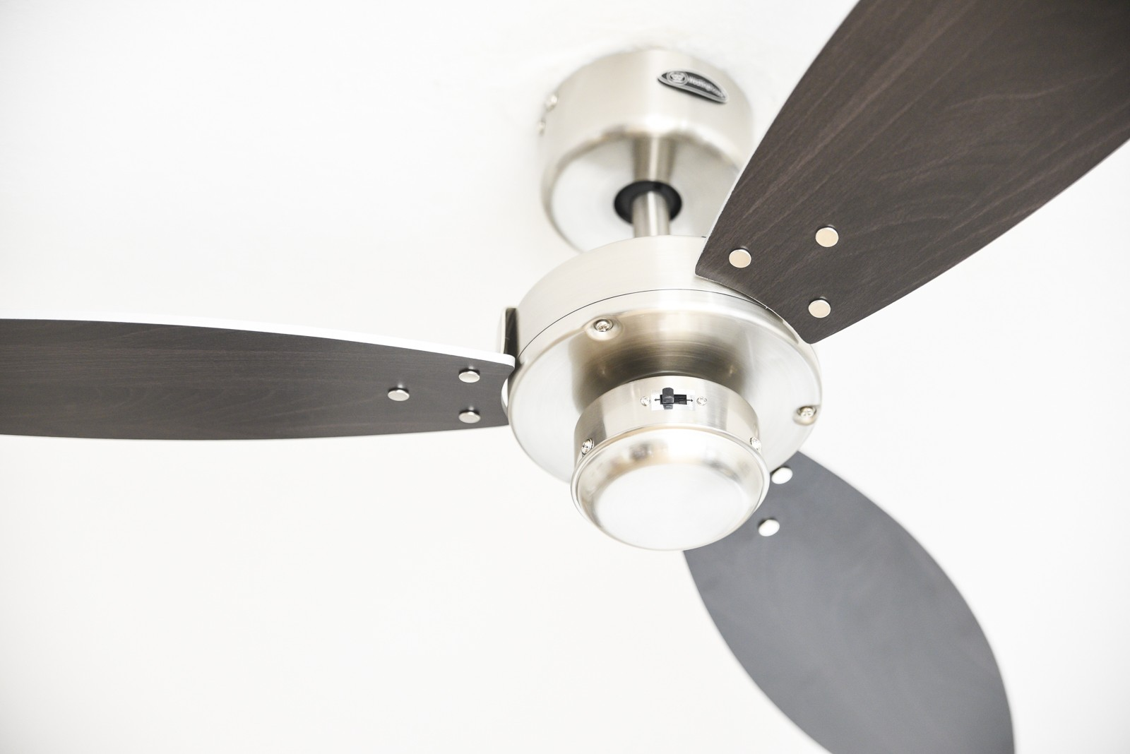 Deckenventilator mit drei Flügeln, silbernem Motor und dunkelgrauen Flügeln. Weißer Deckenhintergrund.
