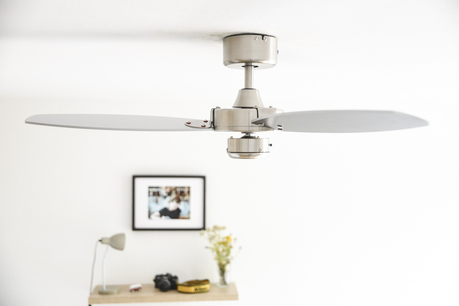 Silberner Deckenventilator. Verschwommener Vordergrund mit Bild, Blumen und Lampe auf einem Regal.