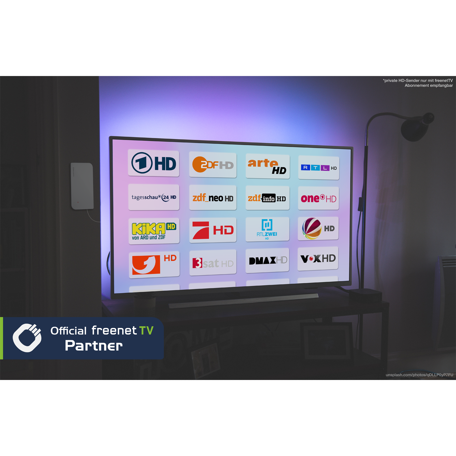 Ein TV-Bildschirm zeigt Senderlogos. Ein schwarzer TV-Ständer, eine Lampe und eine weiße Wandbox sind im Raum sichtbar.