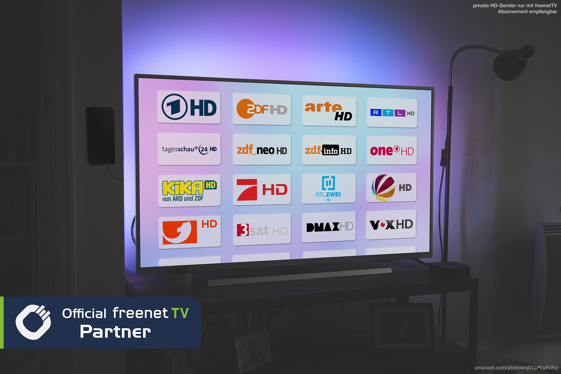 TV-Bildschirm mit Senderlogos. Schwarzer Tisch, Lampe und blaues/lila Licht dahinter.