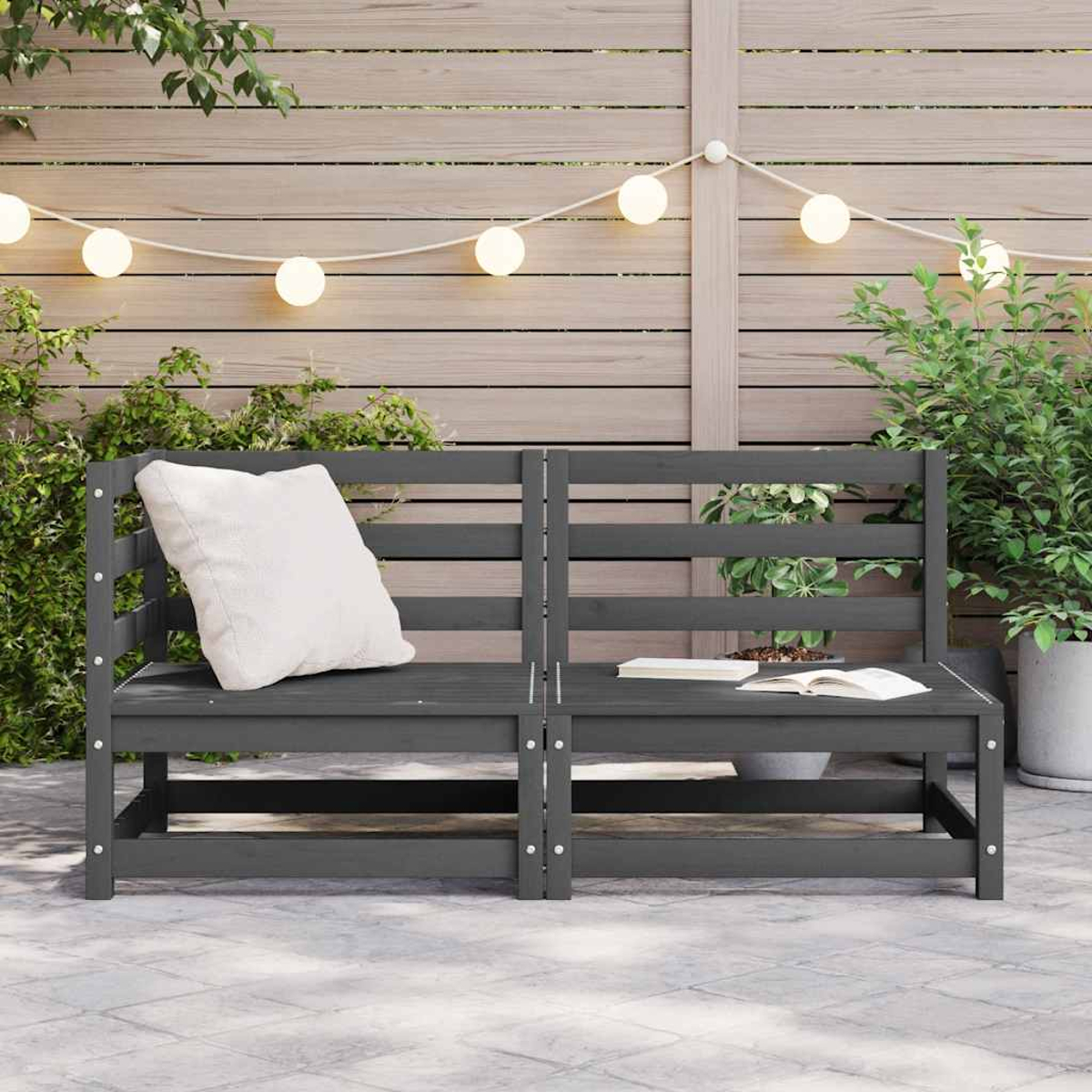 Graue Holzbank mit weißem Kissen und offenem Buch. Garten mit Pflanzen und Lichterketten im Hintergrund.