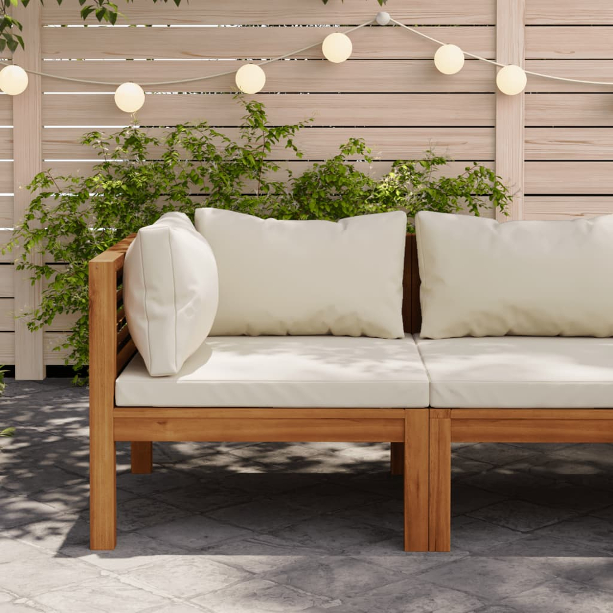 Outdoor-Sofa mit beigen Kissen. Holzrahmen, grüne Pflanzen und weiße Lichter im Hintergrund.