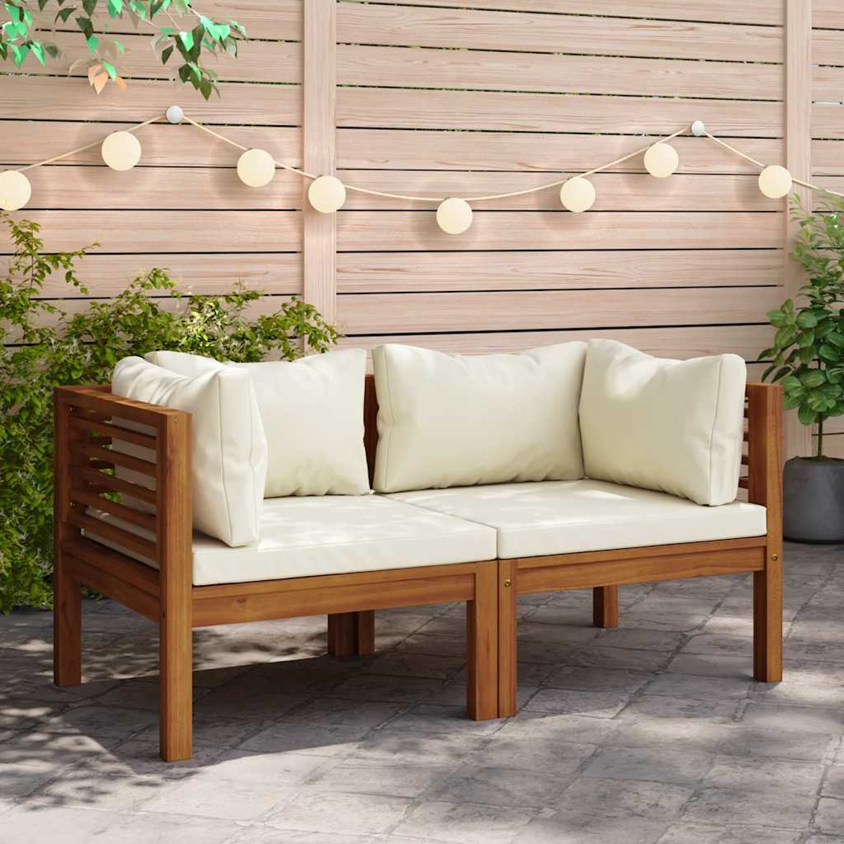 Zwei hölzerne Outdoor-Sofas mit cremefarbenen Kissen. Eine Lichterkette und ein Holzzaun sind im Hintergrund.