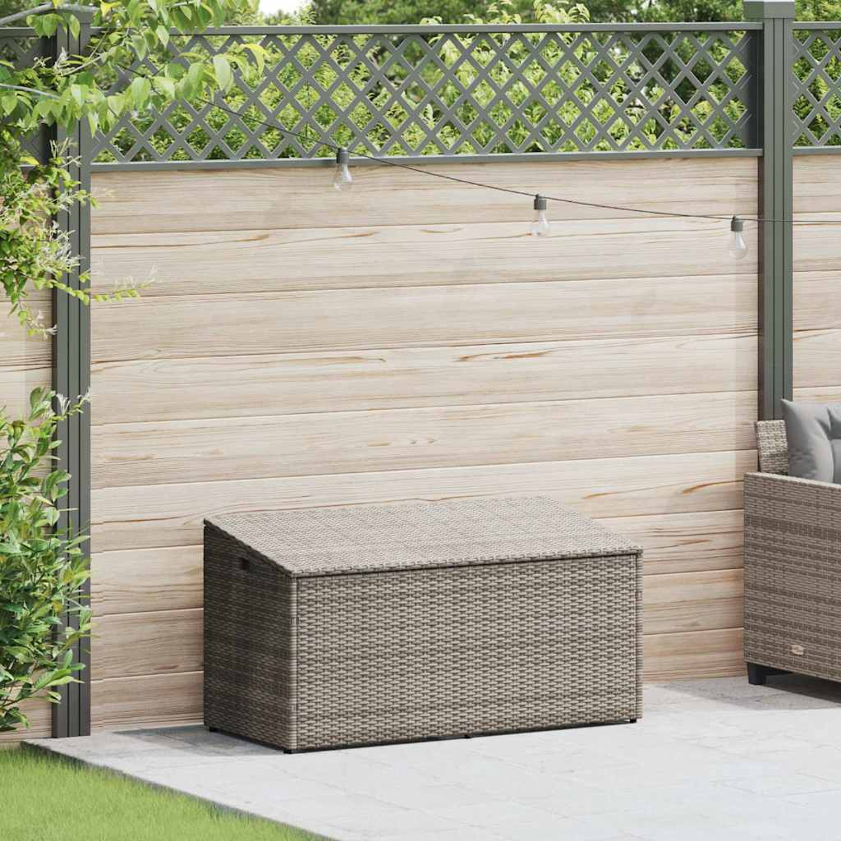Graue Rattan-Aufbewahrungsbox in einem Garten. Box mit geschlossenem Deckel, grauer Zaun und helle Holzwand.