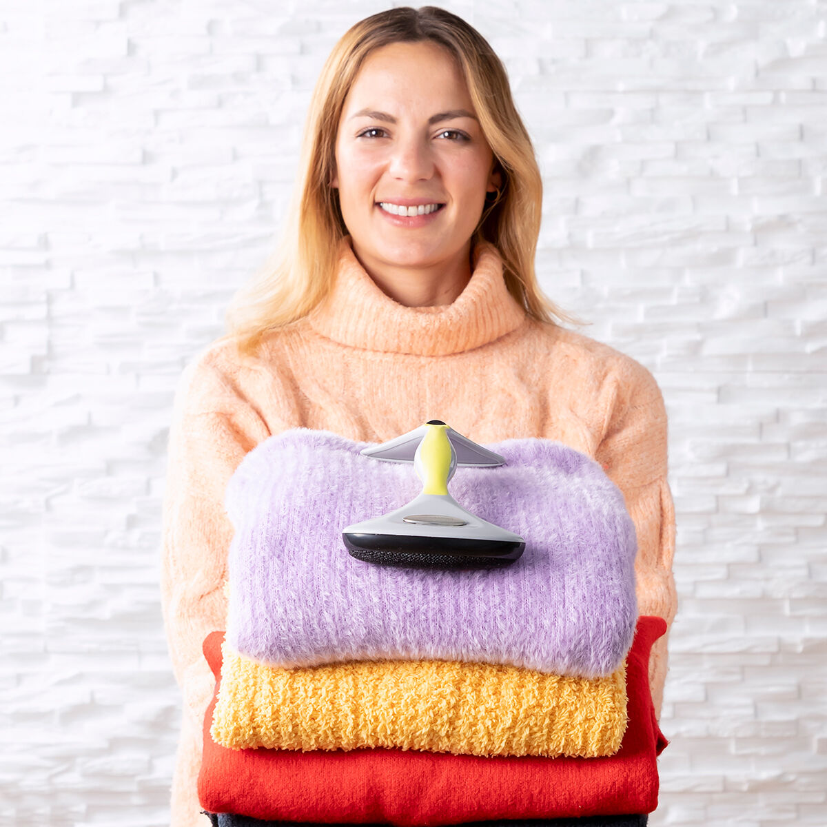 Eine Frau hält gefaltete Pullover mit einem Fusselentferner vor einer Ziegelwand.
