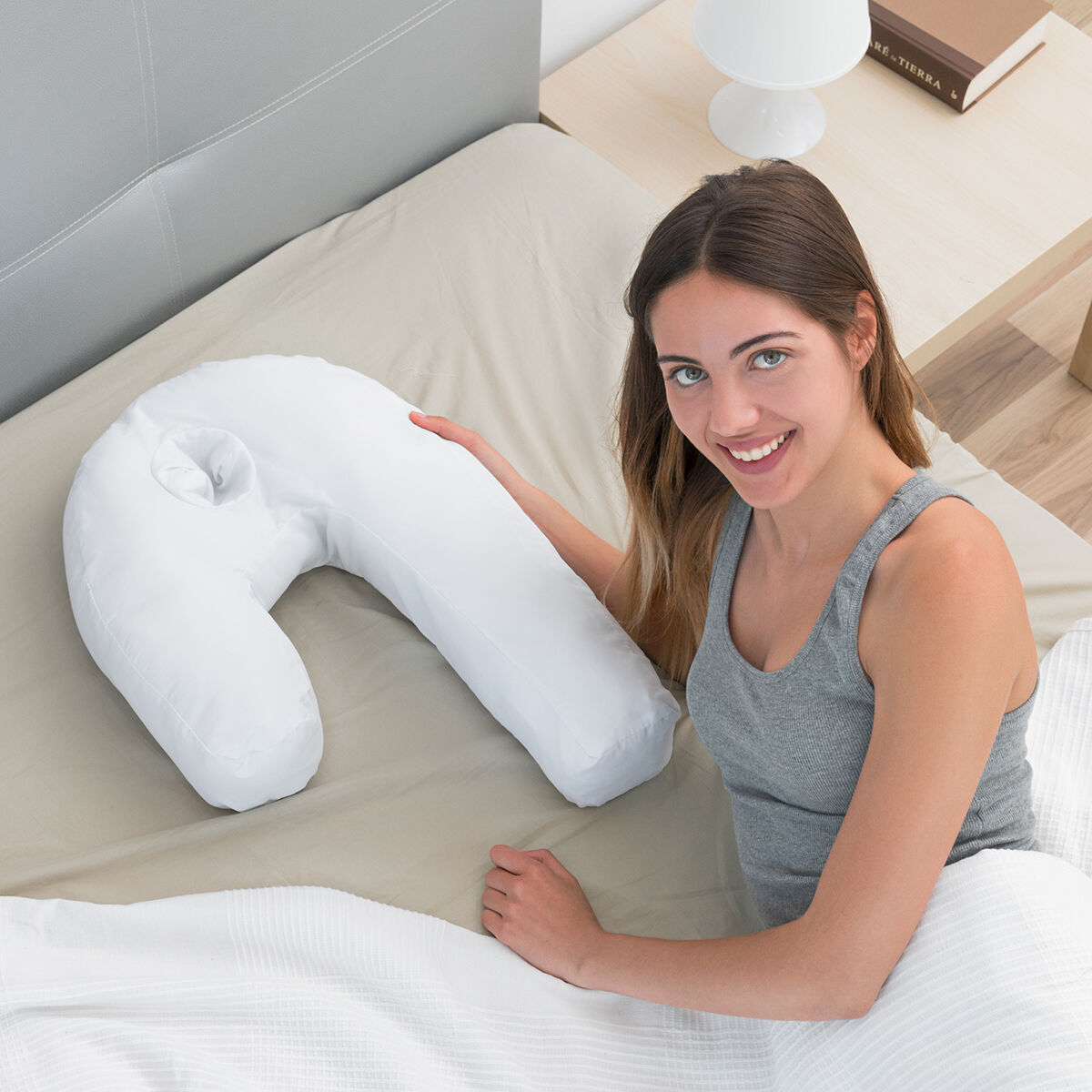 Eine Frau lächelt neben einem weißen U-förmigen Kissen auf einem Bett mit Nachttisch und Buch.