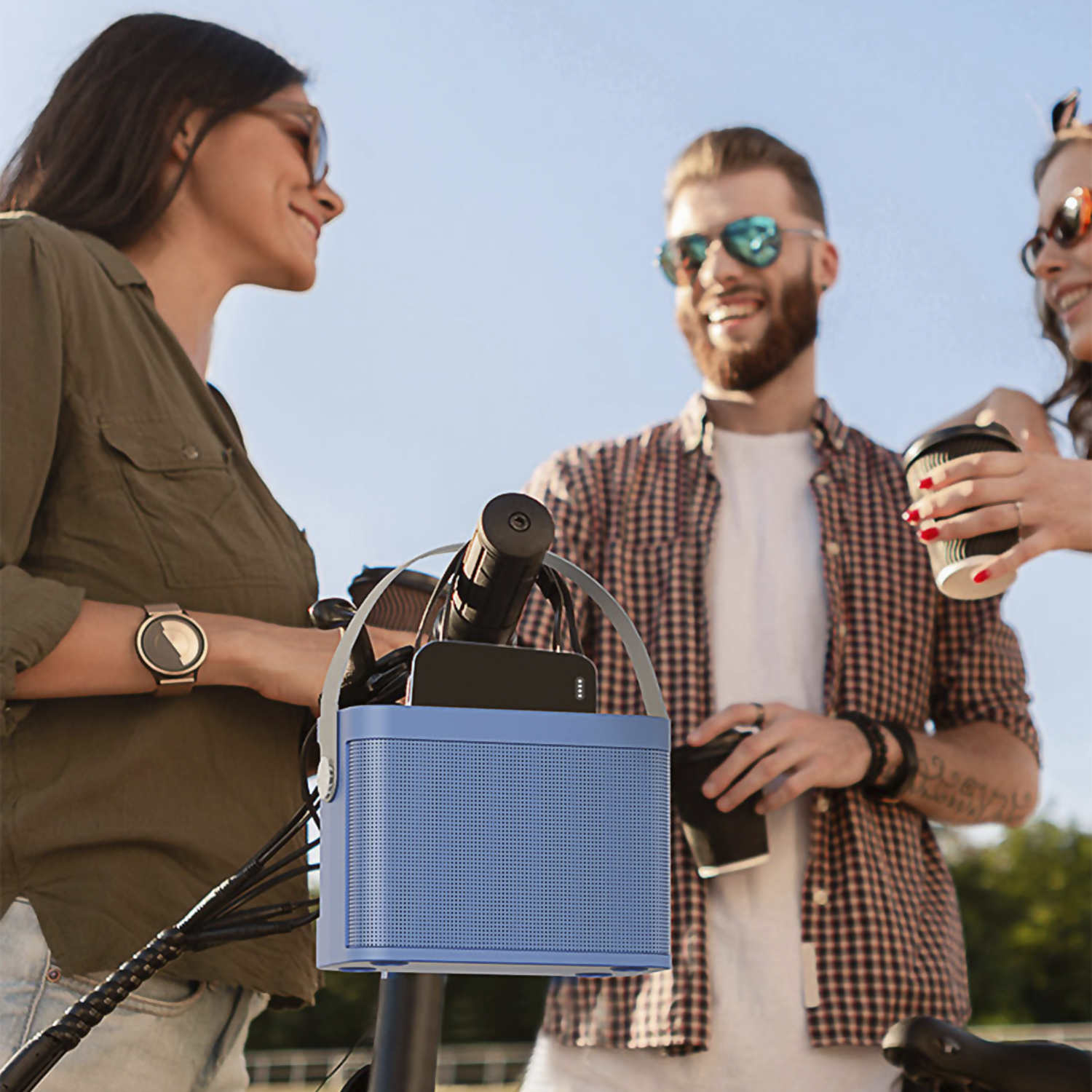 Tres personas con gafas de sol, dos sosteniendo tazas, una con un altavoz azul en una bici.