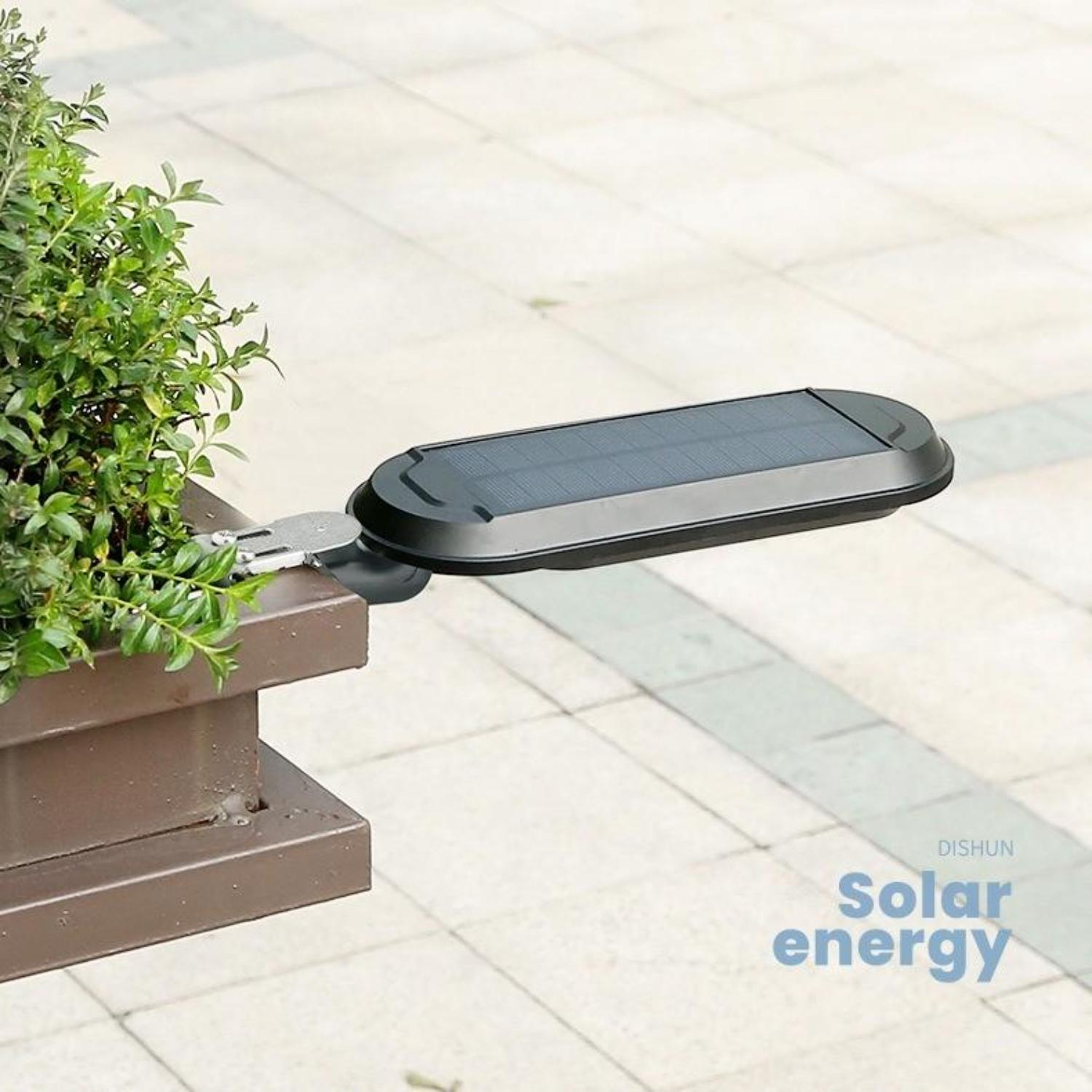 Ein schwarzes Solarpanel ist an einem braunen Pfosten mit grünen Pflanzen befestigt. Der Boden ist hellgrau.
