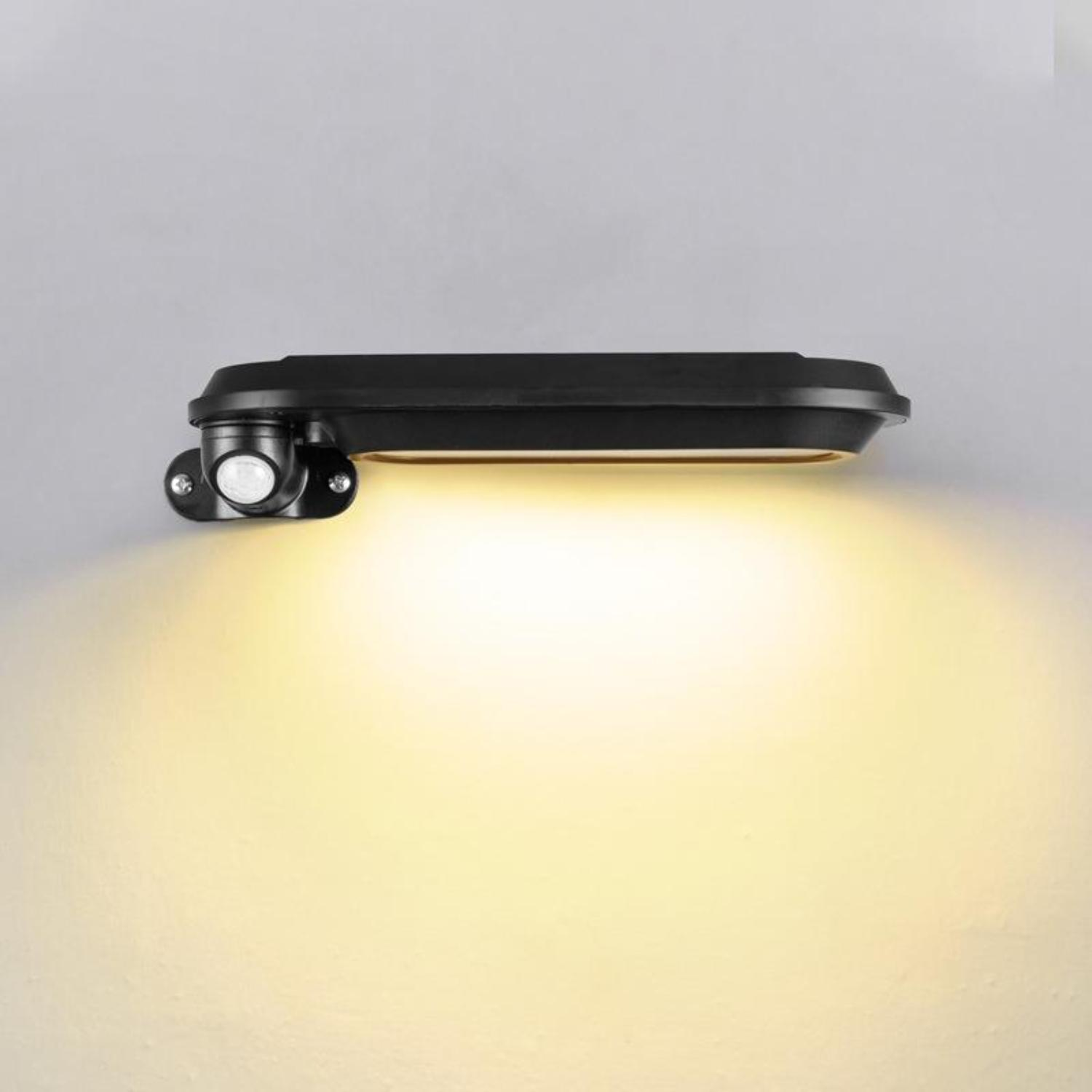 Eine schwarze Außenleuchte an einer grauen Wand. Sie strahlt ein warmweißes Licht aus.