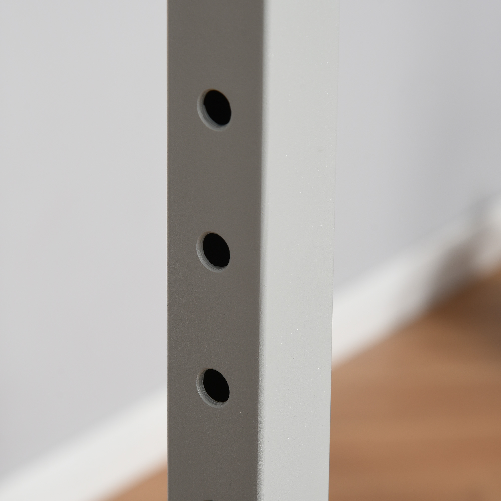 Primer plano de una barra de metal gris con tres agujeros en una pared blanca con piso de madera en el fondo.