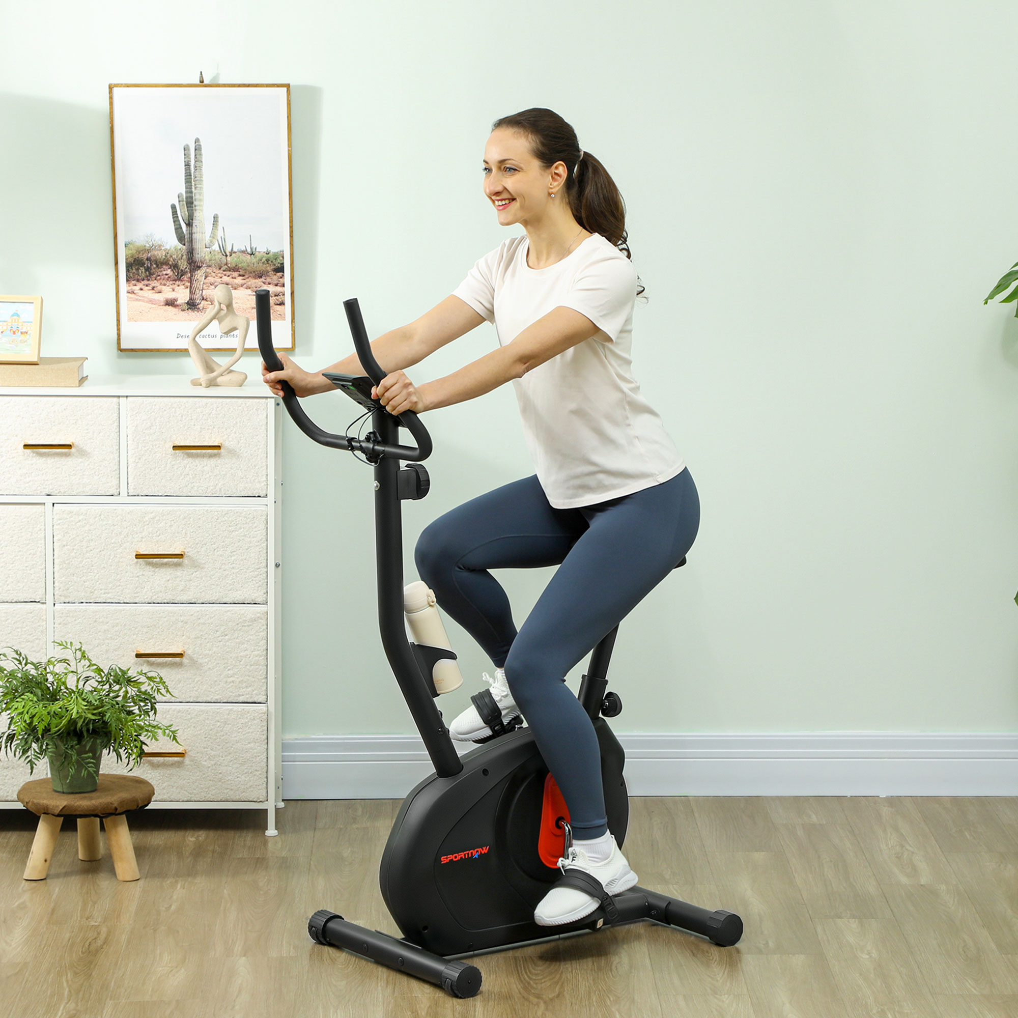 Mujer en una bicicleta estática, sonriendo, en una habitación con una imagen de cactus y un armario blanco.
