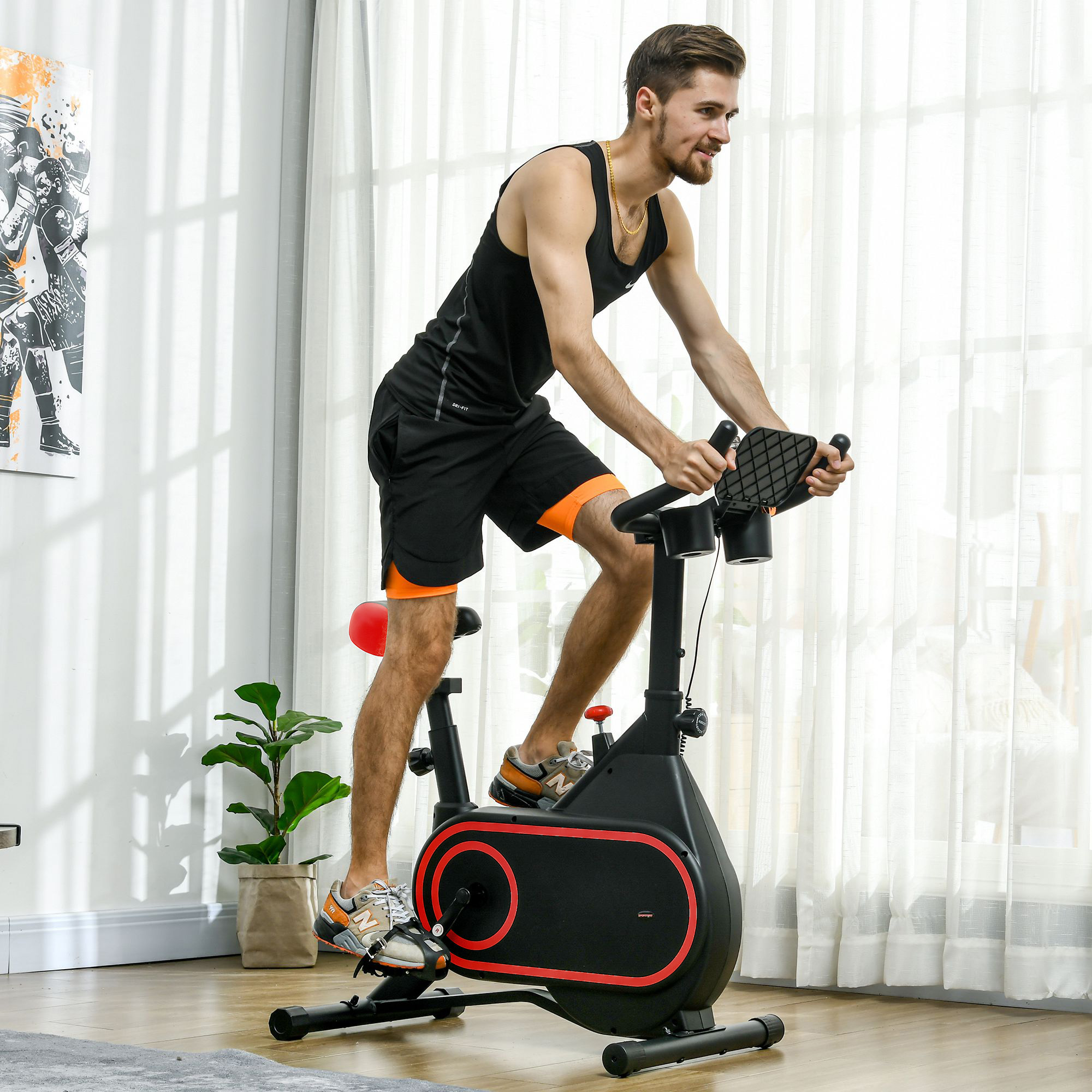 Hombre en una bicicleta estática negra y roja en una habitación con una planta, un cuadro y una ventana.