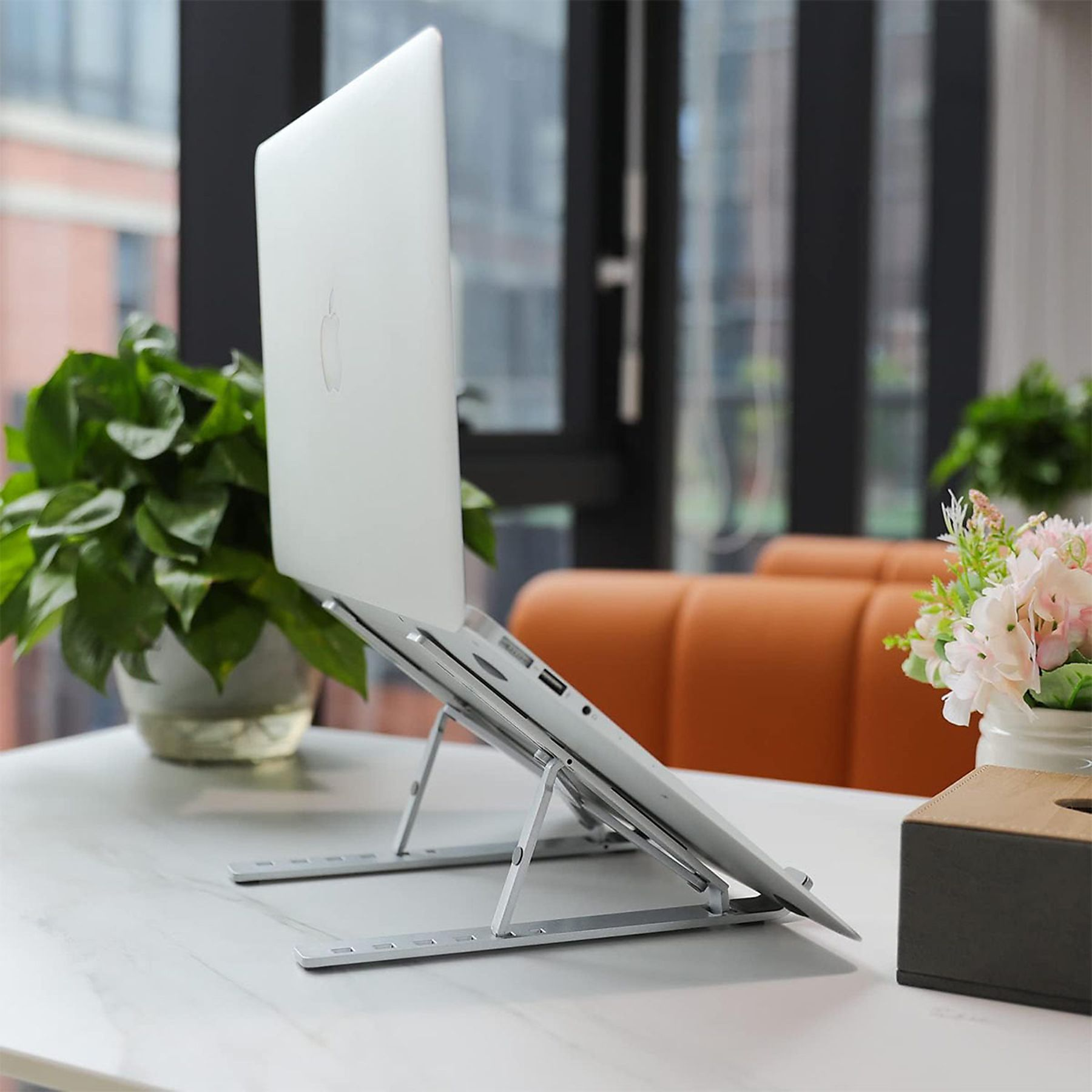 Laptop auf einem silbernen Ständer auf einem Tisch. Eine Pflanze und eine Vase sind ebenfalls sichtbar.