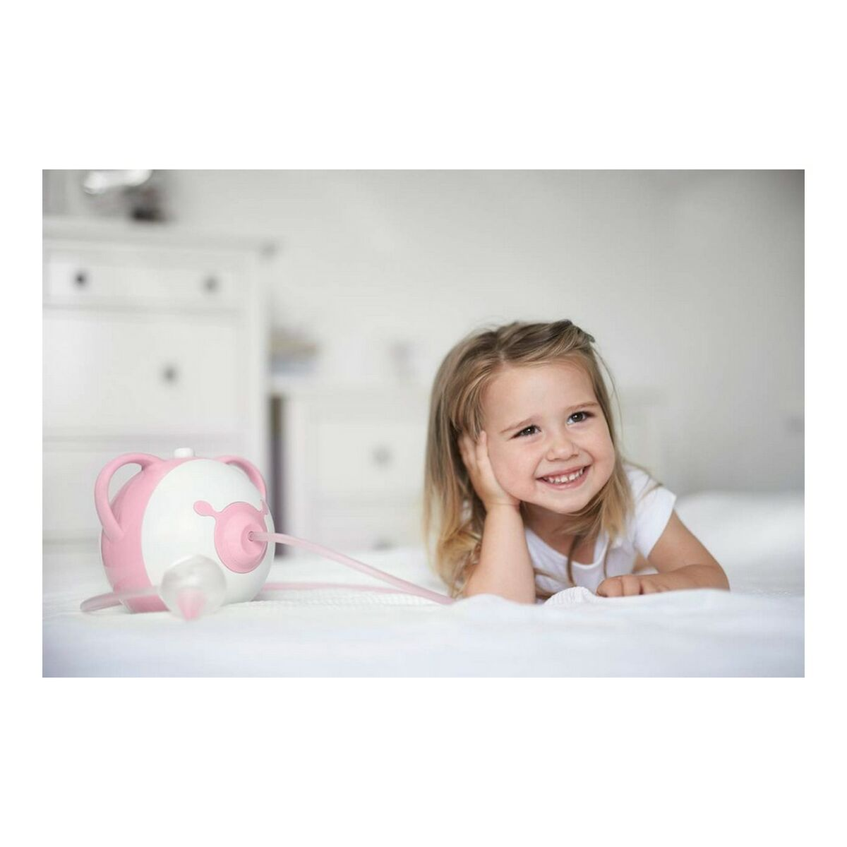 Ein lächelndes Kind liegt auf einem Bett und betrachtet einen rosa-weißen Nasensauger.