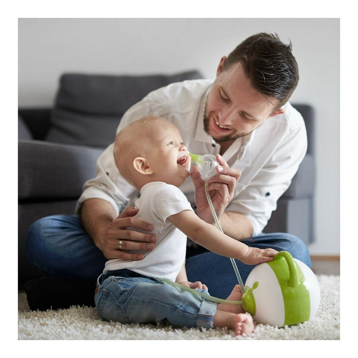 Ein Baby inhaliert aus einem grünen Inhalator, gehalten von einem lächelnden Mann. Auf Teppich.