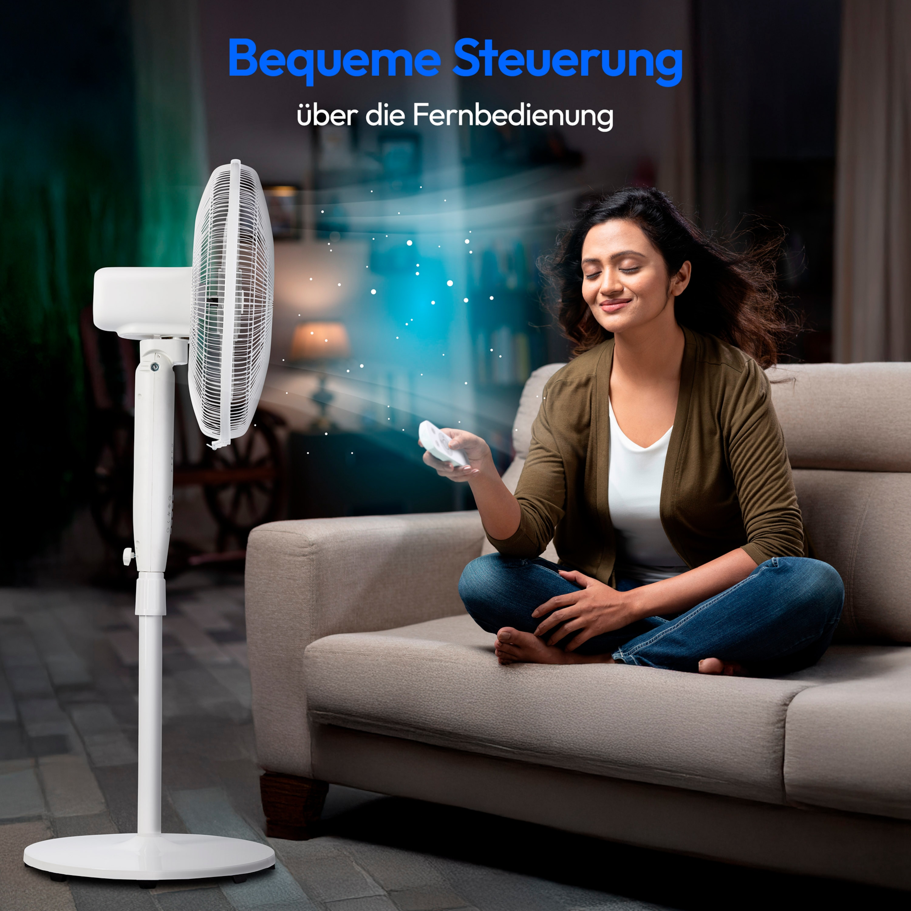 Eine Frau sitzt auf einem Sofa und bedient eine Fernbedienung. Ein weißer Ventilator steht daneben.