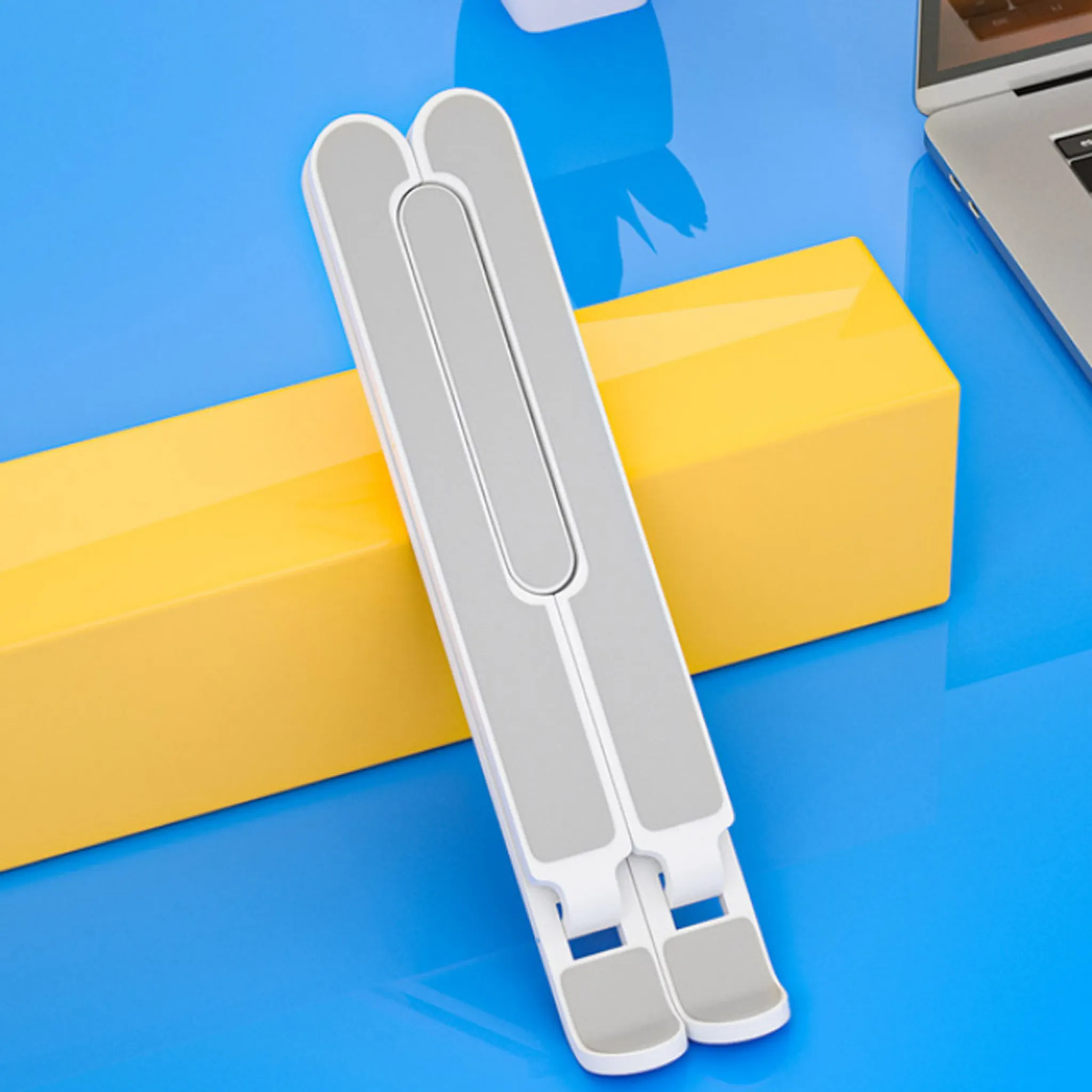 Zusammengeklappter Laptopständer, auf blauer Oberfläche platziert. Der Ständer ist weiß und grau.