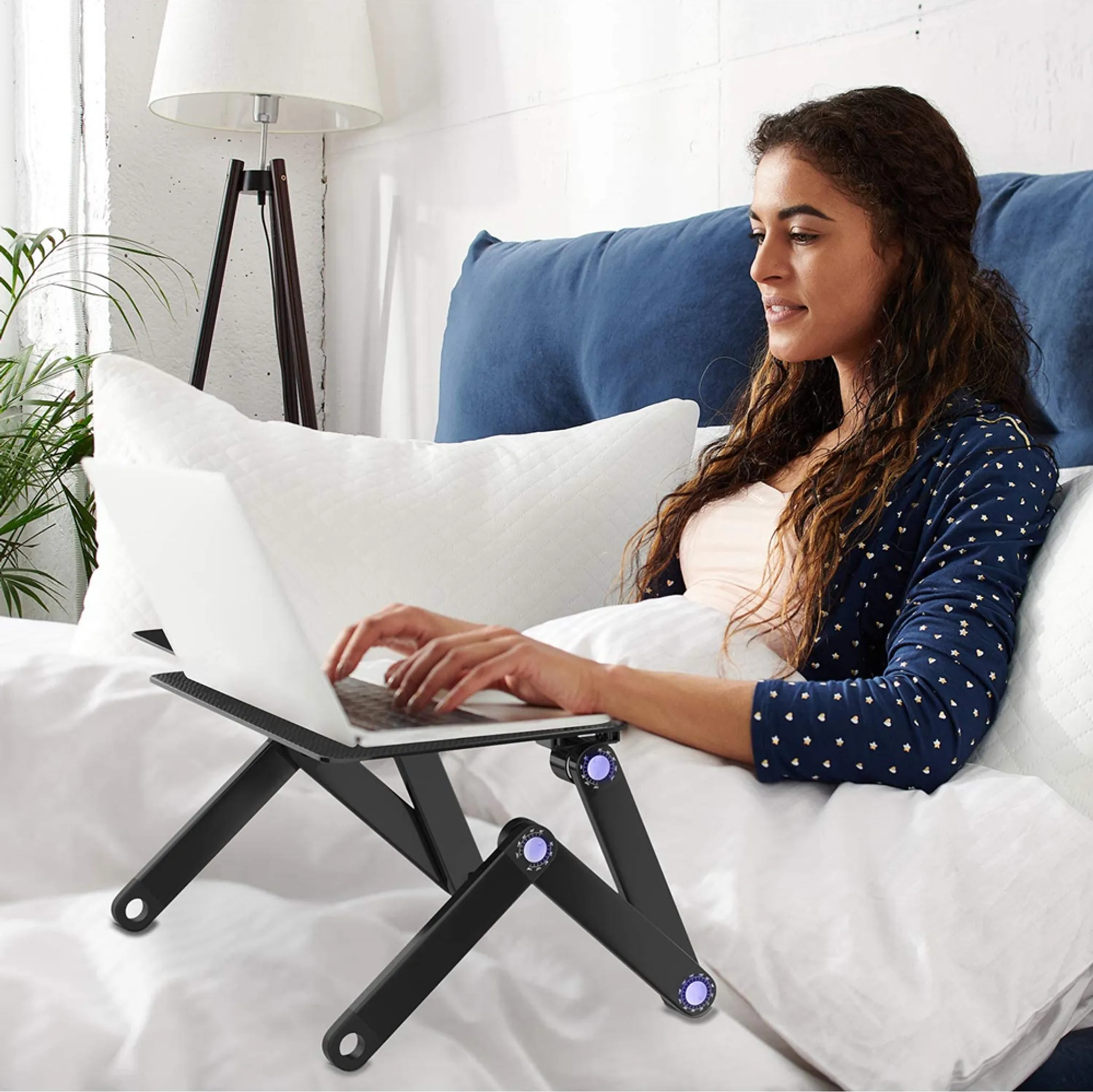 Frau im Bett benutzt Laptop auf verstellbarem schwarzen Laptopständer, Lampe in der Nähe.