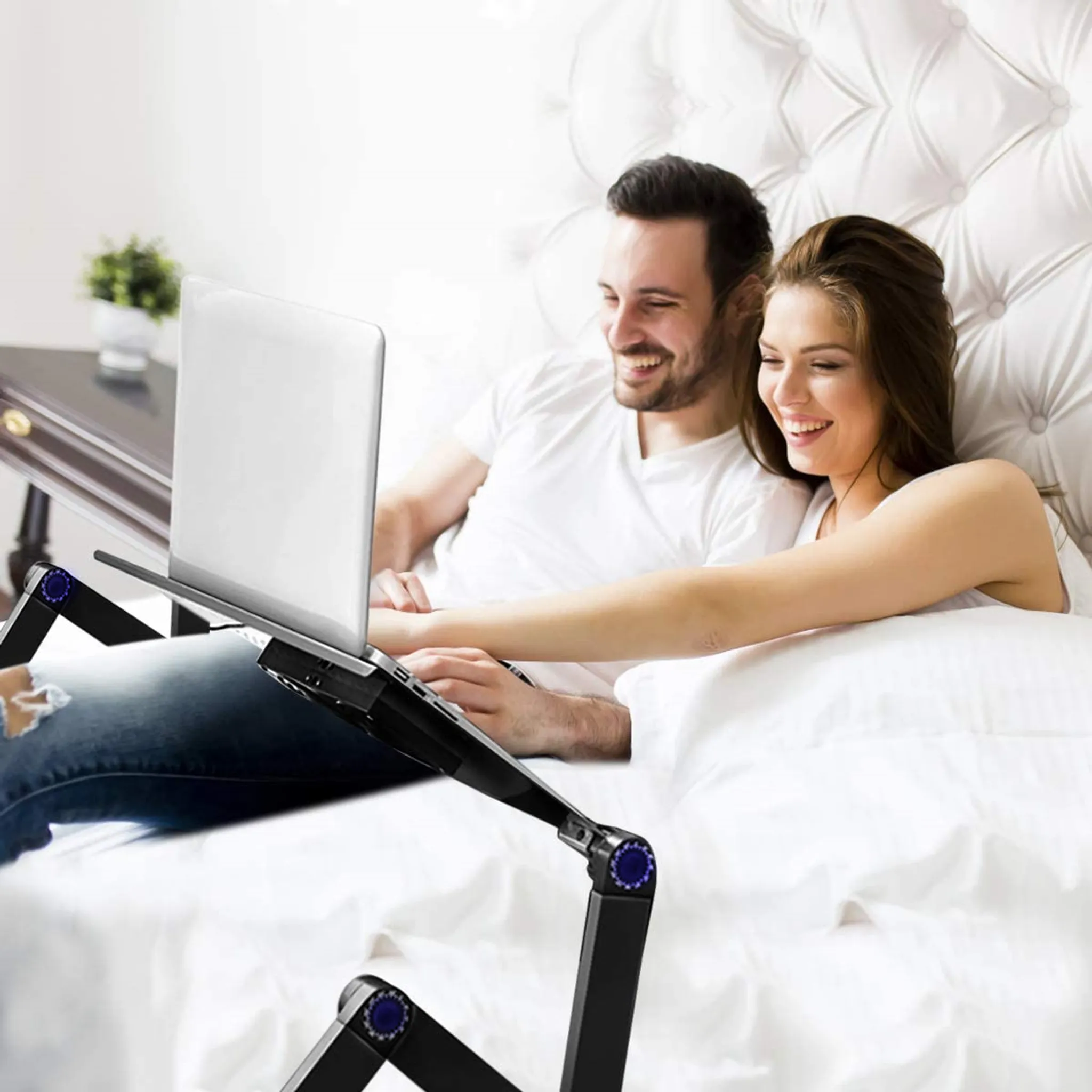 Paar benutzt Laptop auf verstellbarem Schreibtisch im Bett, lächelnd, im Schlafzimmer.