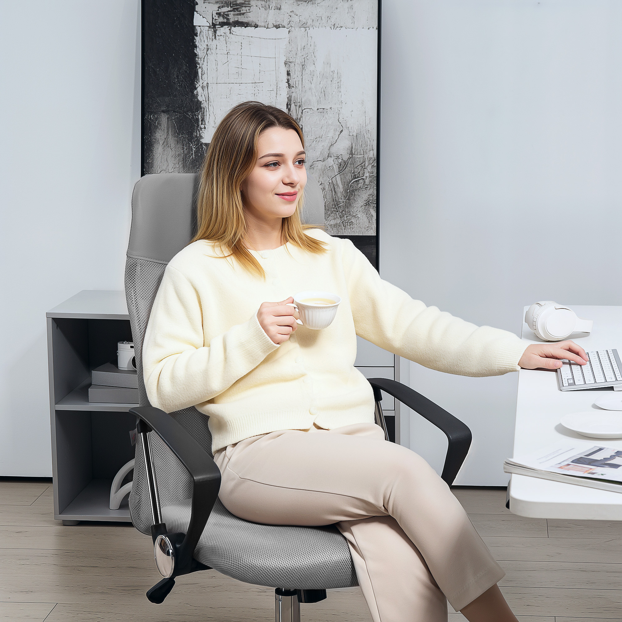 Una mujer con un suéter crema está sentada en un escritorio, bebiendo café y usando un teclado, mirando al lado.