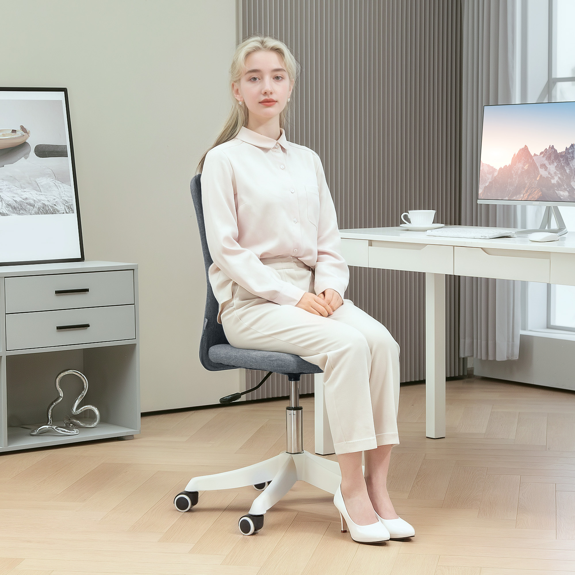 Mujer sentada en una silla de oficina gris, frente a una computadora, con zapatos y pantalón blancos.