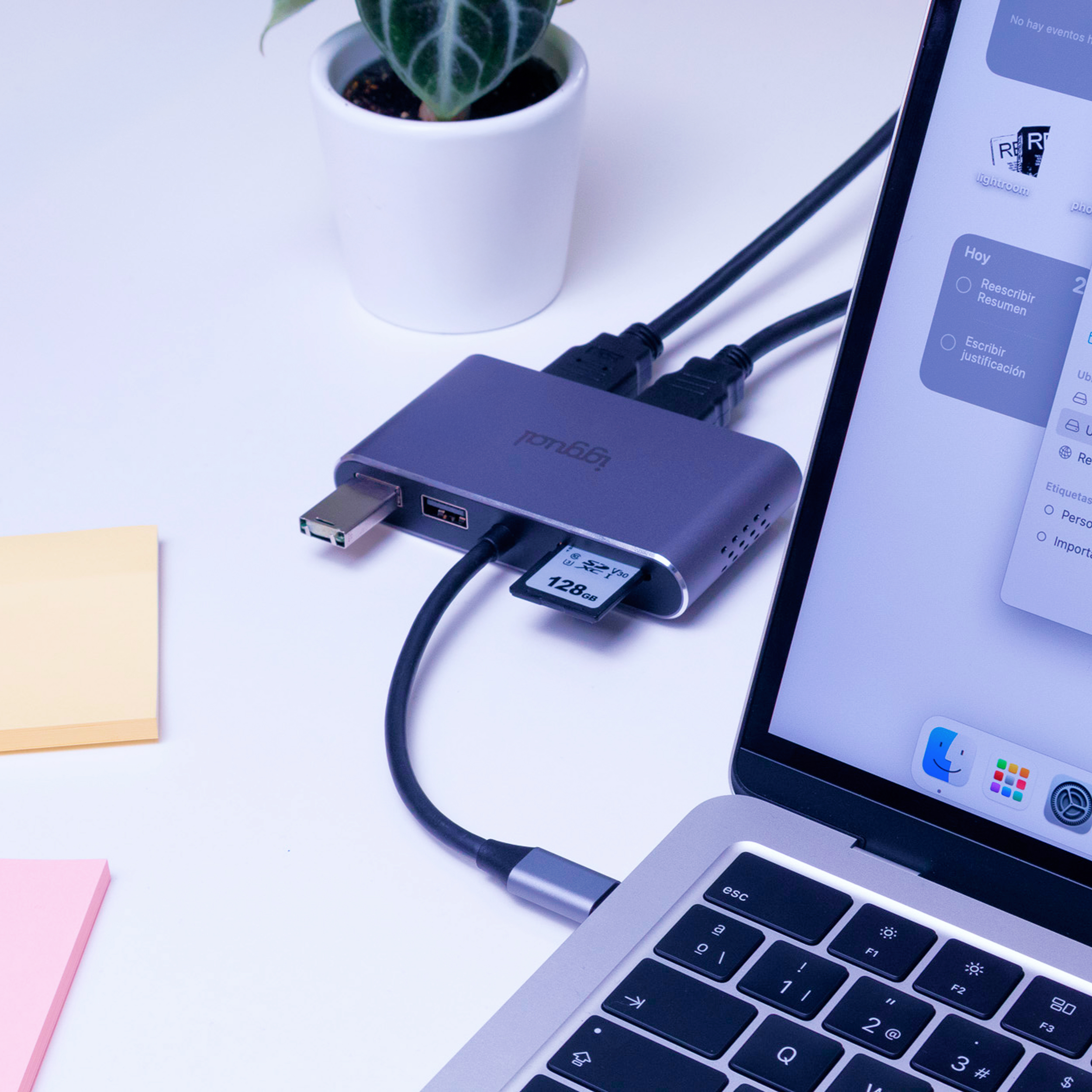 Laptop podłączony do adaptera z portami USB, HDMI i kartą SD. Obok roślina i karteczki samoprzylepne.