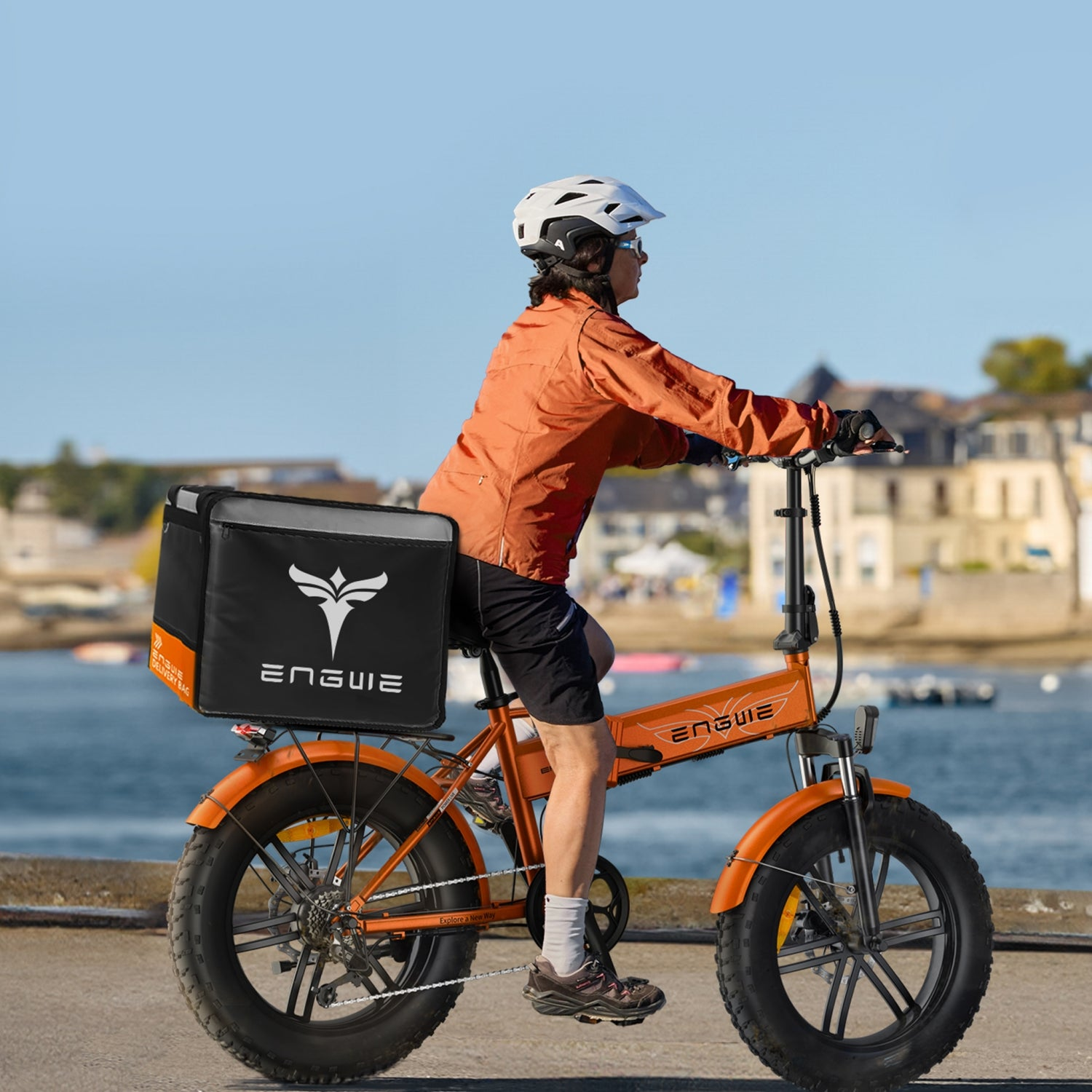 Una persona montando en bicicleta eléctrica naranja con carga. El fondo tiene agua y edificios.