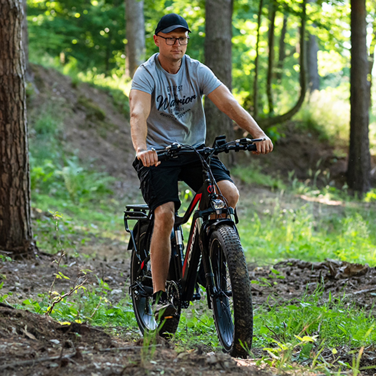 Mann fährt ein schwarzes und rotes Fatbike auf einem Waldweg, trägt Mütze und Brille.