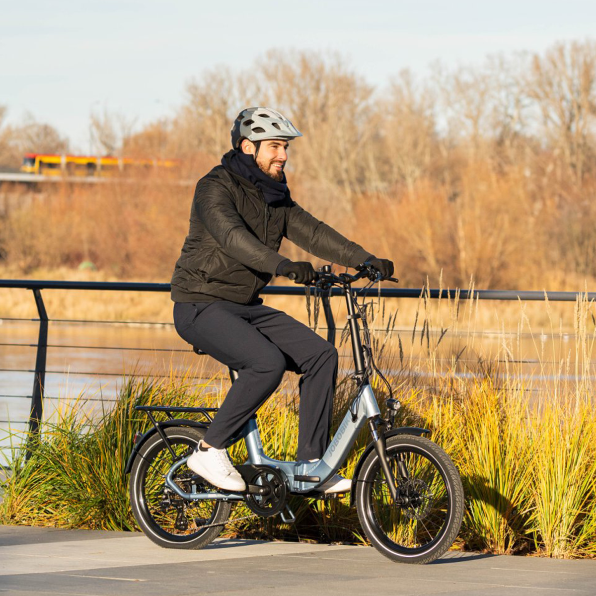 Mężczyzna jadący na rowerze elektrycznym. Ma na sobie kask i uśmiecha się. Rower ma bagażnik.