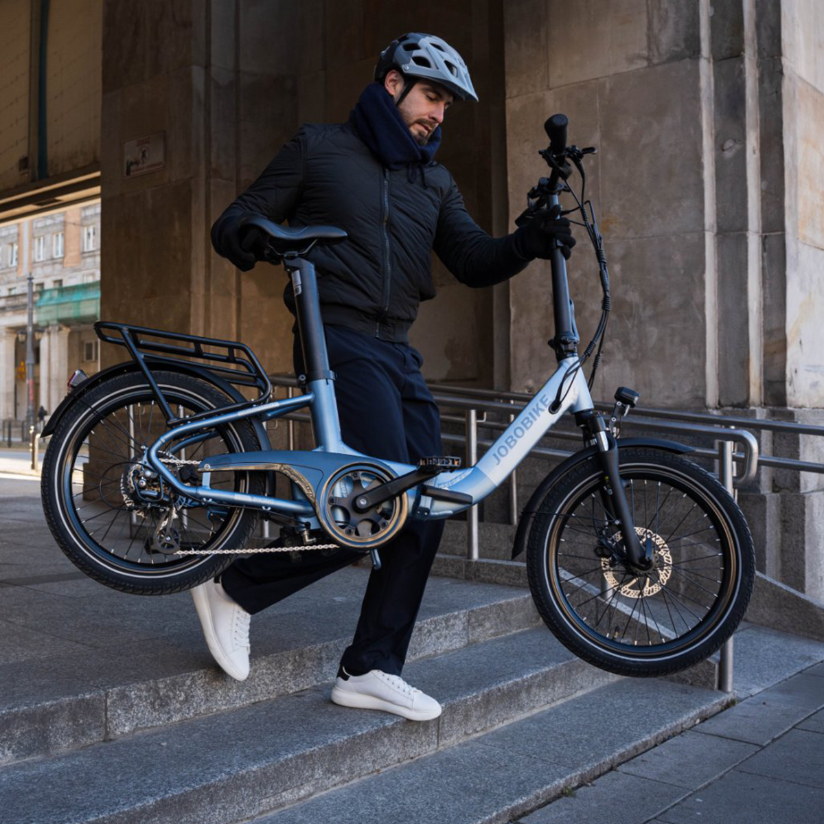 Mężczyzna niosący niebieski rower elektryczny JOBOBBIKE po schodach, w kasku.