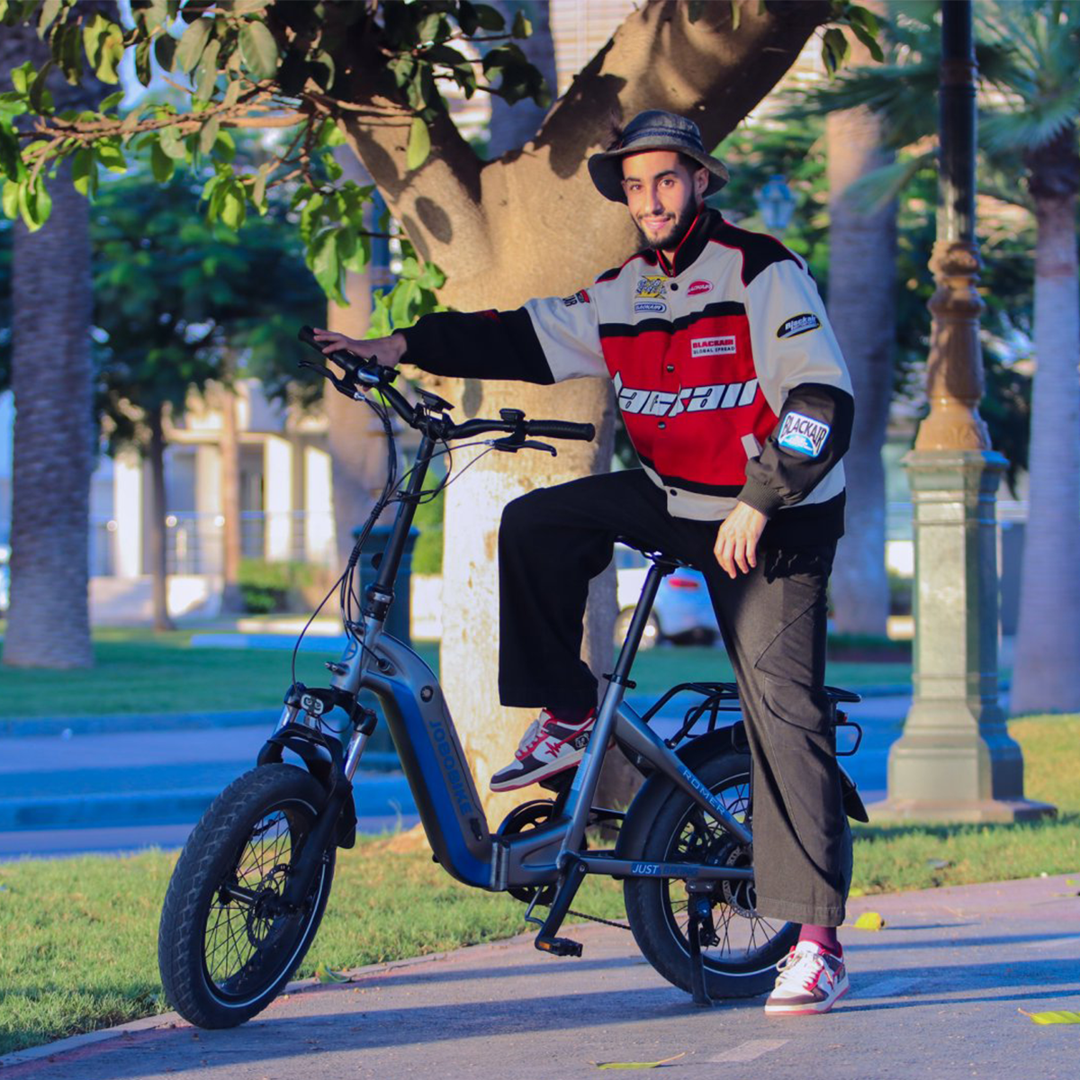 Mężczyzna w kurtce siedzi na szarym rowerze elektrycznym. Rower jest w pobliżu parku.