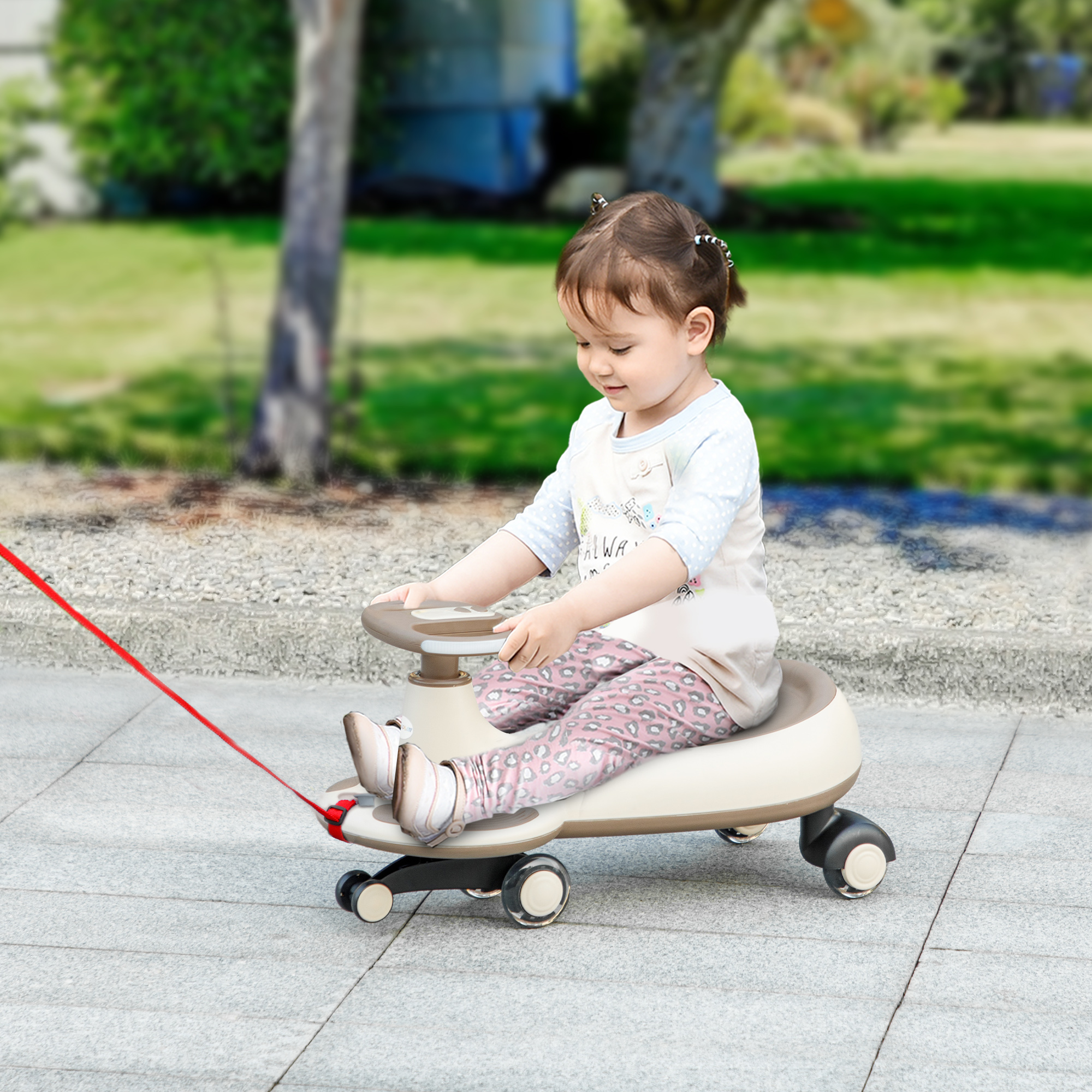 Una niña pequeña con coletas sentada en un coche de juguete beige sobre una superficie pavimentada con un árbol y vegetación al fondo.