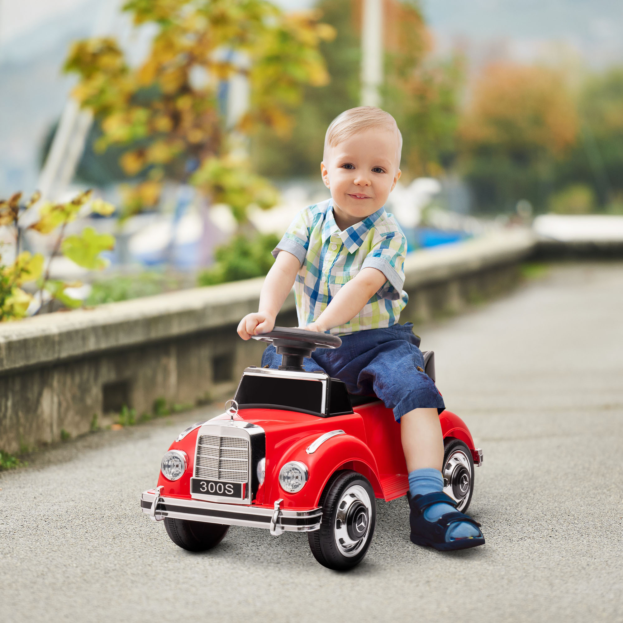 Un niño con camisa azul conduce un coche de juguete rojo por una acera, con árboles y agua al fondo.
