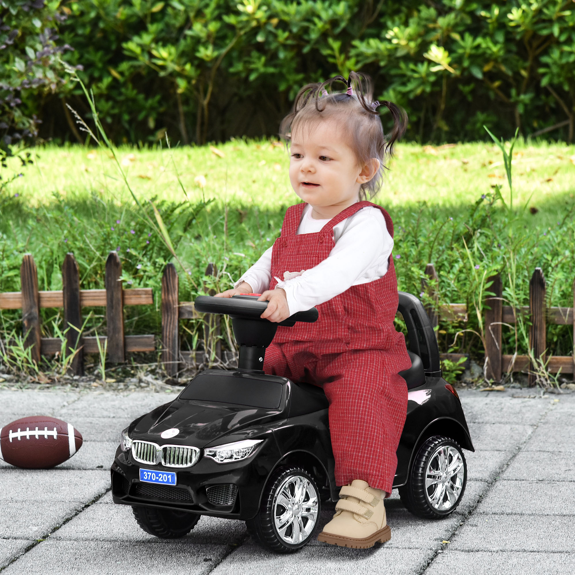 Un bambino con una tuta rossa è seduto su un'auto giocattolo nera all'esterno con un pallone da calcio, davanti a una recinzione di legno.