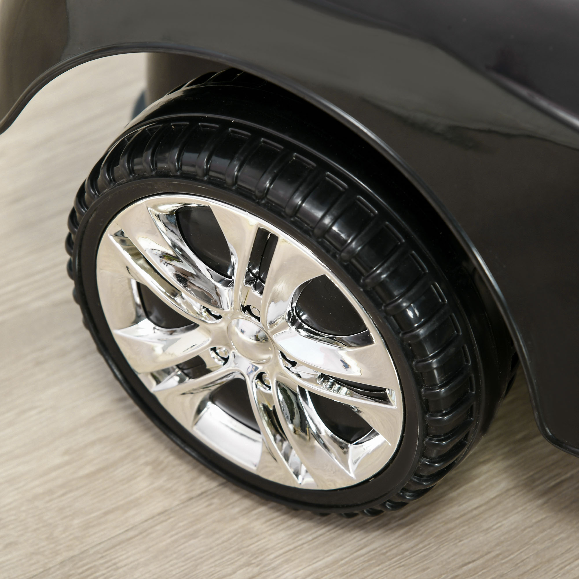 Primer plano de una rueda cromada con un neumático negro sobre un suelo de madera, parte de un coche de juguete negro.