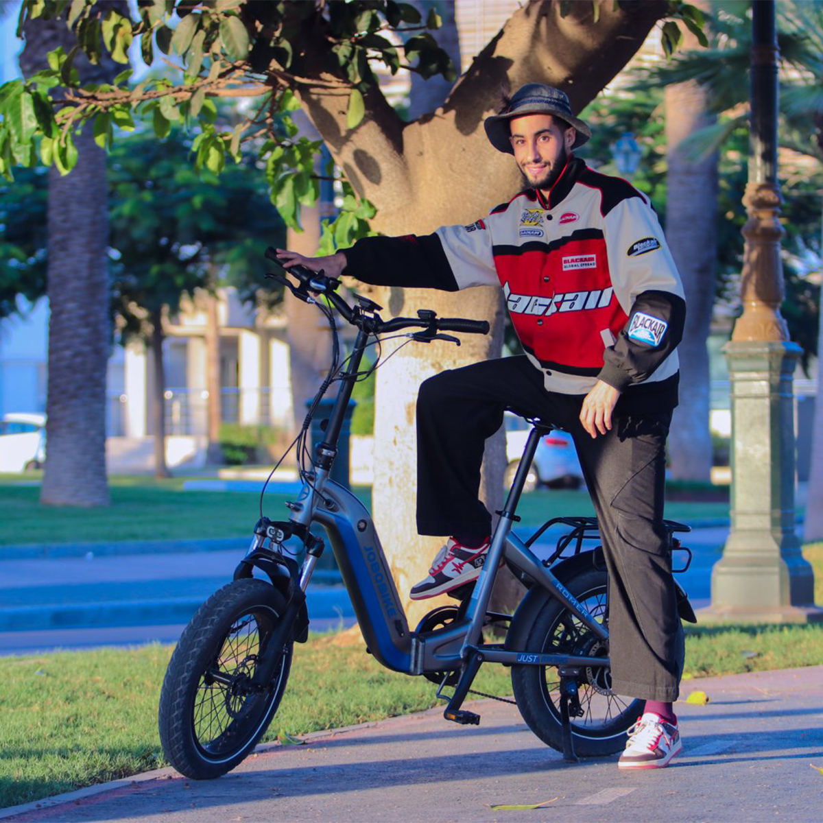 Mężczyzna na szarym rowerze, w kurtce, czapce, okularach przeciwsłonecznych i czarnych spodniach.