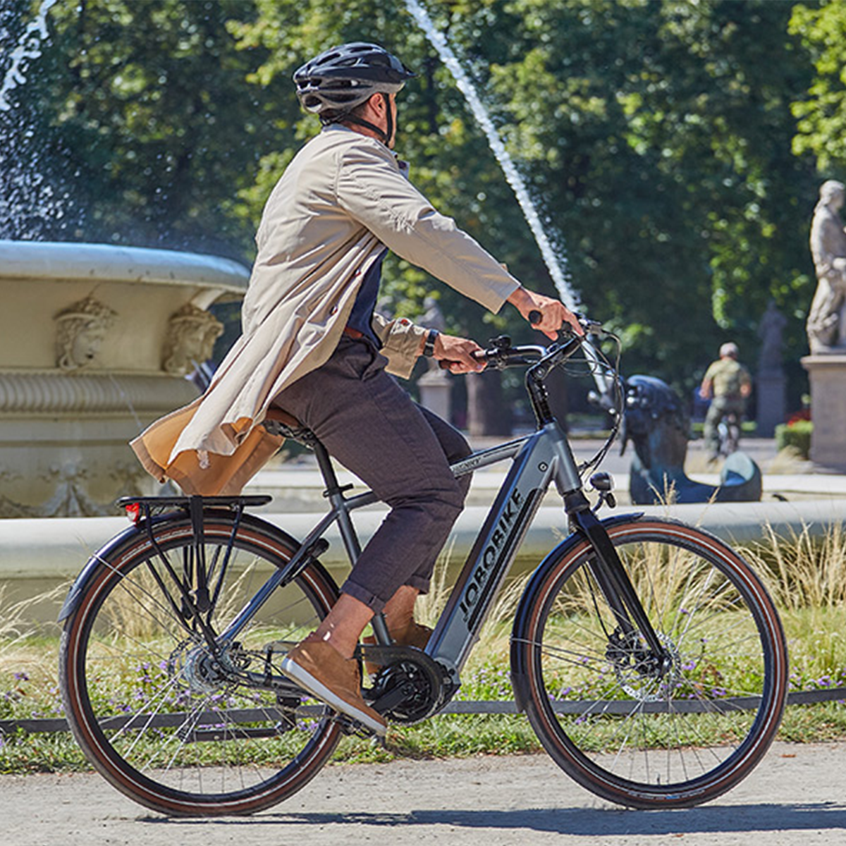 Mężczyzna jedzie szarym JOBOBIKE po ścieżce obok fontanny. Tłem jest park.