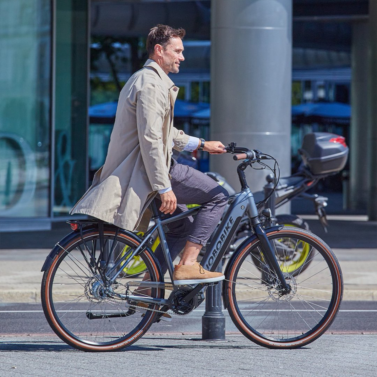 Mężczyzna jadący szarym rowerem elektrycznym na ulicy. Rower marki JOBOBIKE. Beżowy płaszcz, szare spodnie.