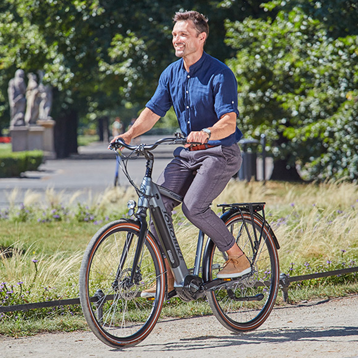 Mężczyzna na szarym rowerze elektrycznym. Zielone drzewa w tle.