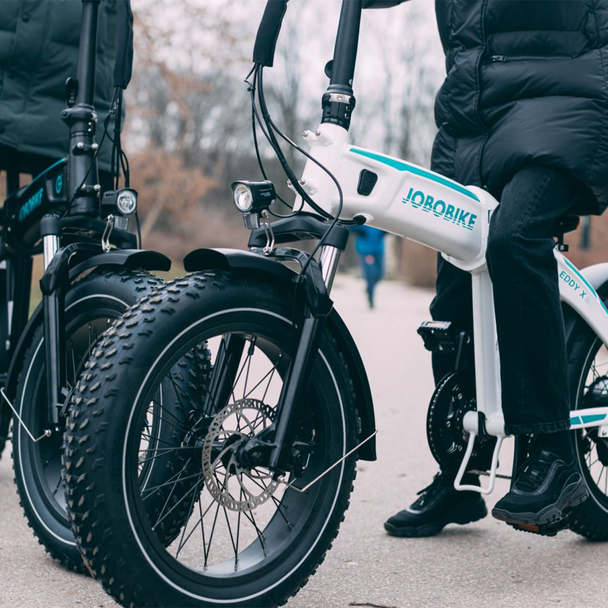 Dwóch mężczyzn jadących rowerami elektrycznymi z dużymi oponami, obok siebie na ścieżce, jeden w czarnej kurtce.