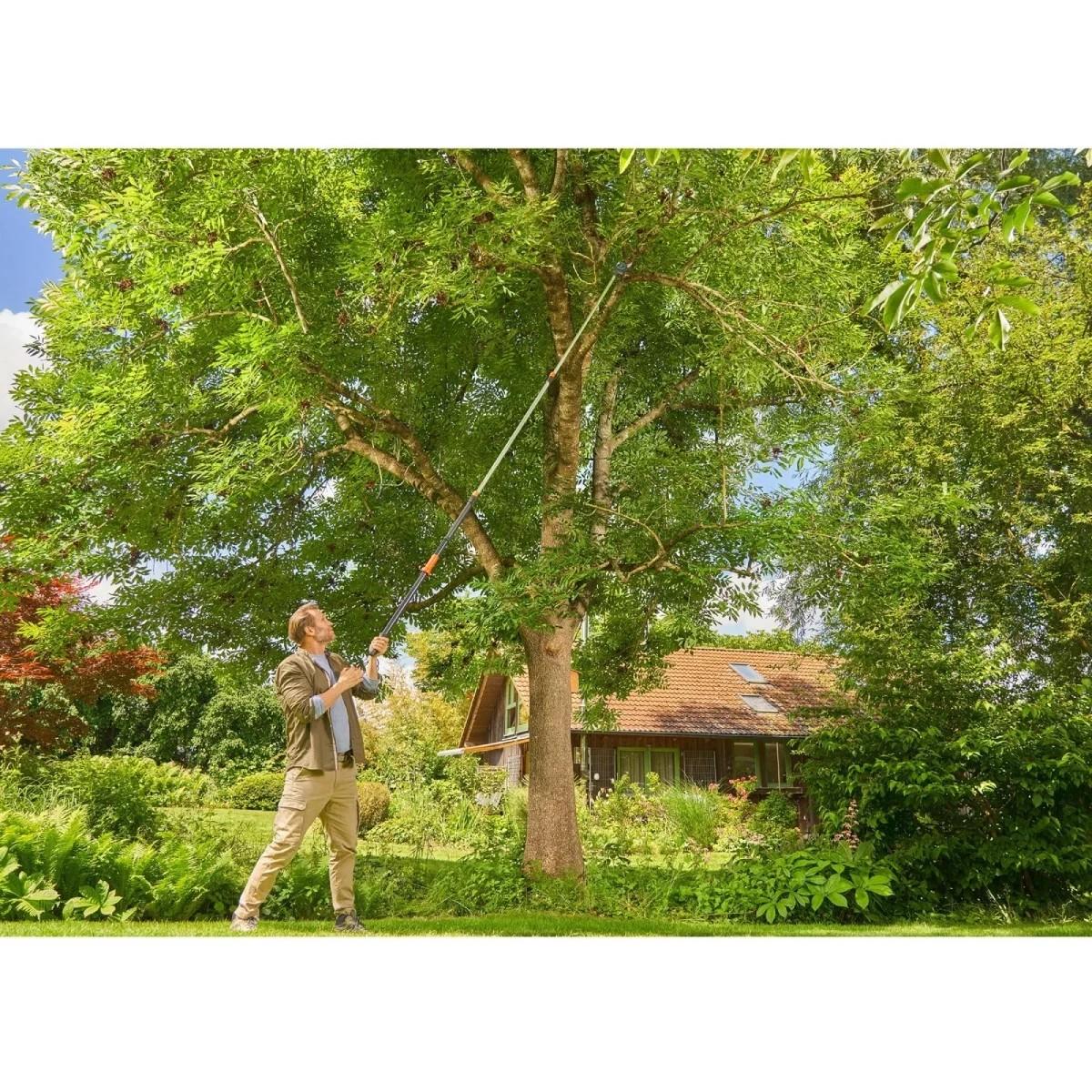 Mężczyzna przycina drzewo sekatorem na długim wysięgniku. Zielona trawa i dom w tle.