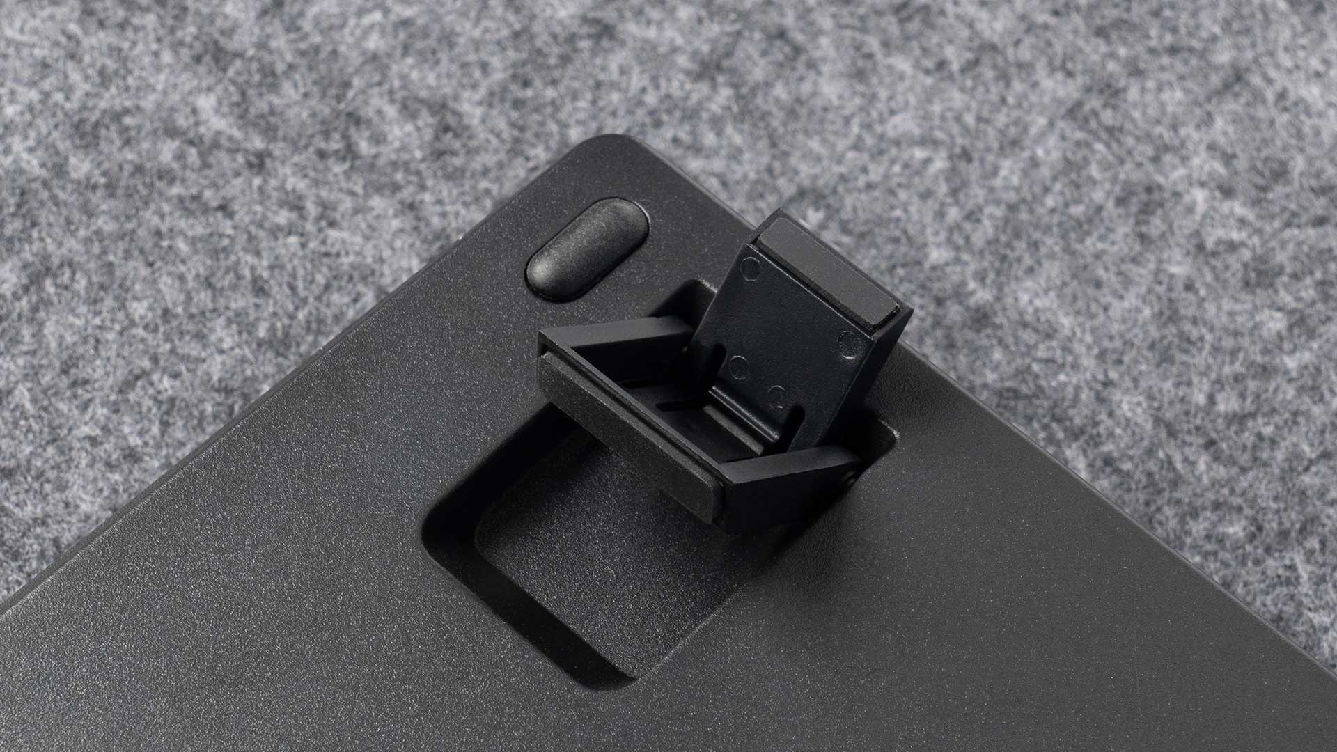 Czarna klawiatura z otwartą przegrodą. Posiada port do połączeń. Klawiatura leży na szarej powierzchni.