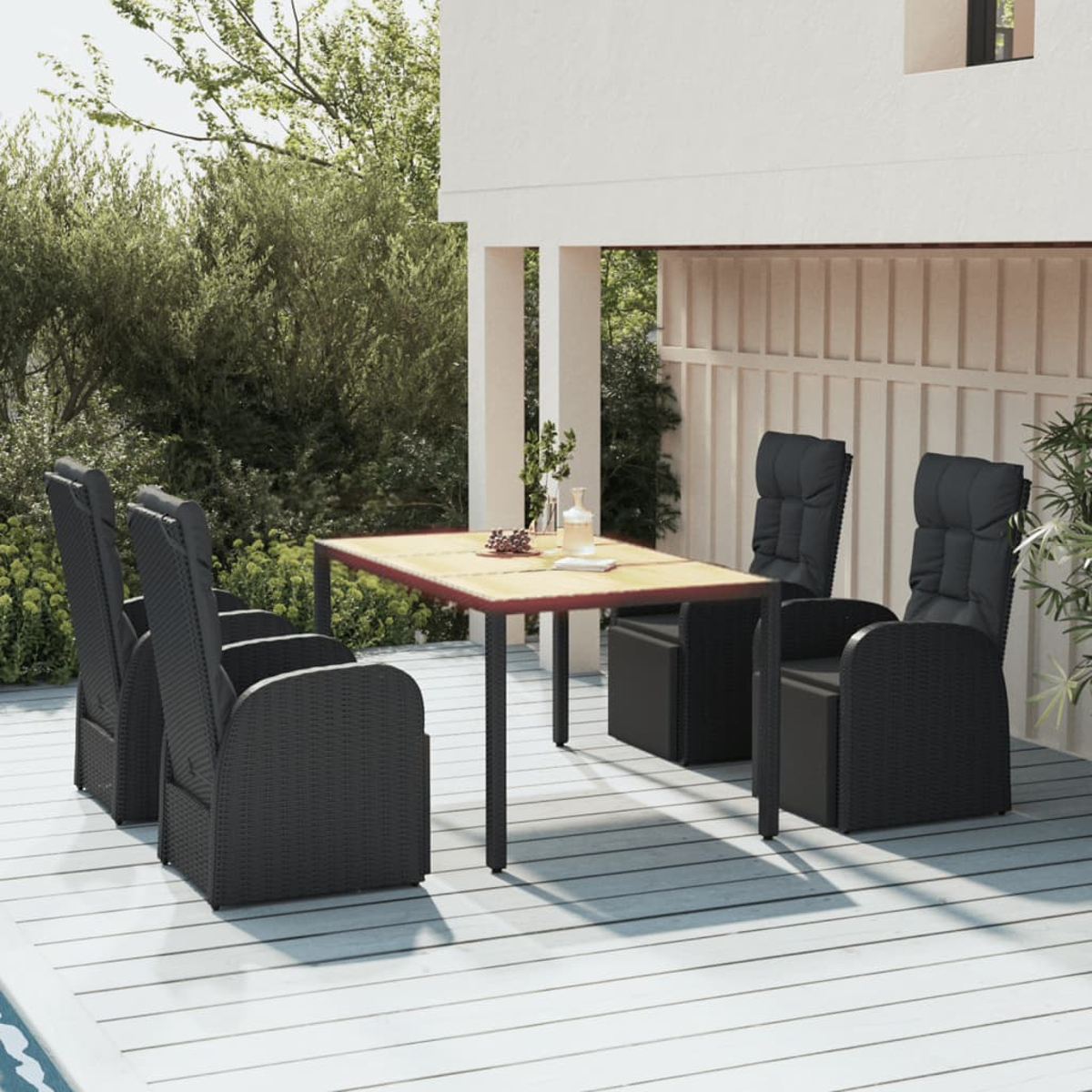 Szene im Freien: Tisch mit Stühlen auf einer Holzterrasse, eine weiße Wand und Bäume im Hintergrund.