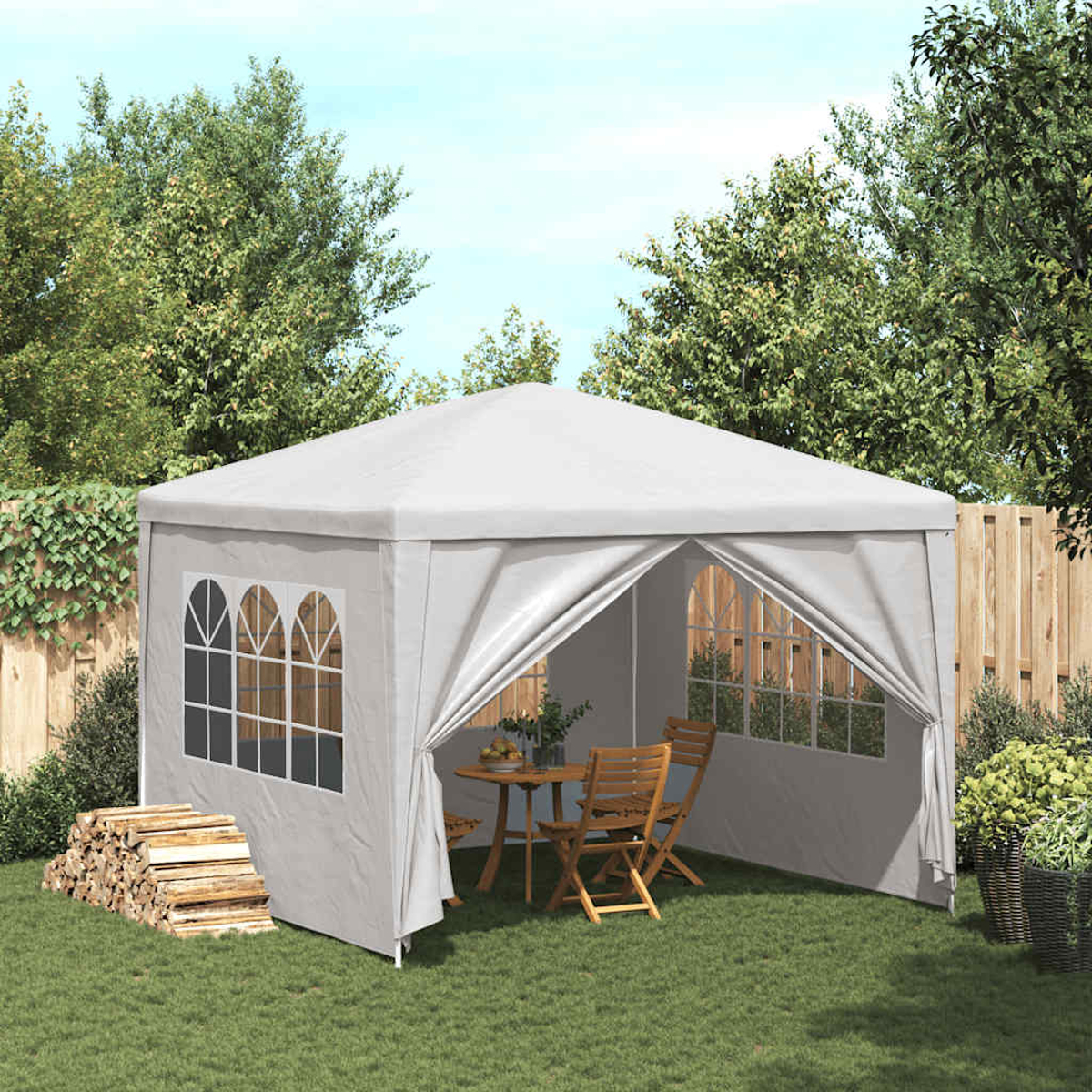 Weißer Pavillon auf Gras. Tisch und Stühle sind drinnen. Holzstapel und Topfpflanzen in der Nähe. Grüne Bäume.