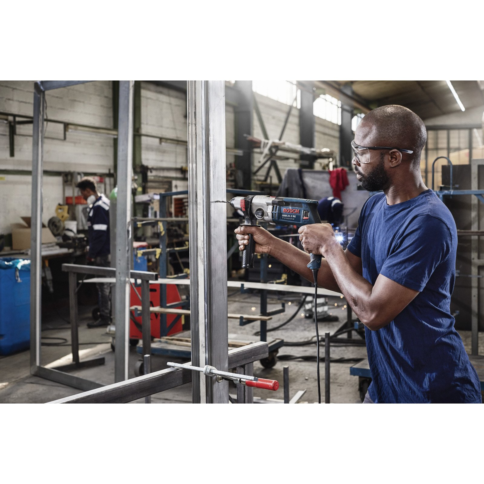 Mężczyzna w niebieskiej koszuli wierci metalową ramę. Inny mężczyzna jest w tle. Ustawienia warsztatu.