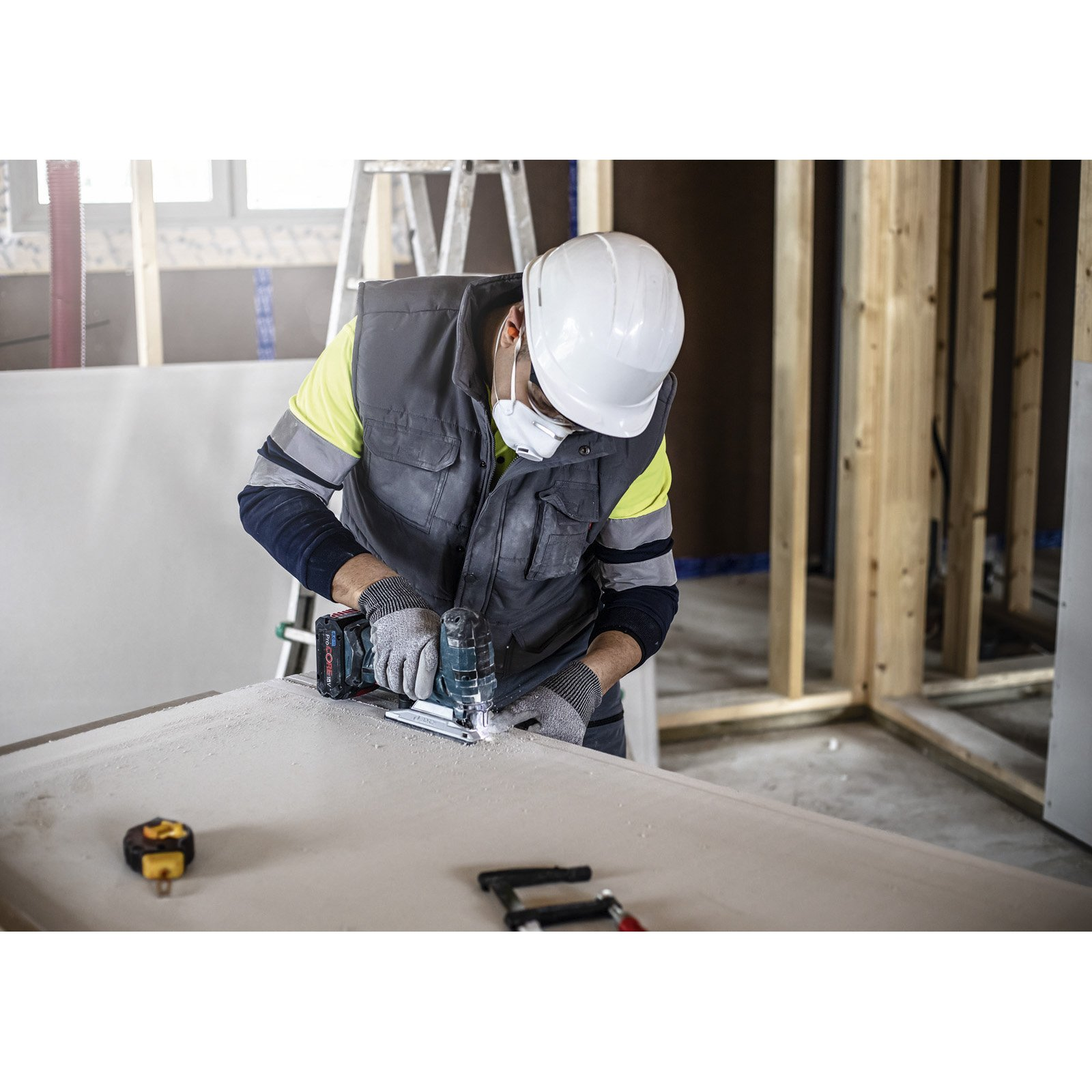 Osoba w masce i kamizelce odcina płyty gipsowo-kartonowe multi-narzędziem. Pył unosi się w powietrzu.