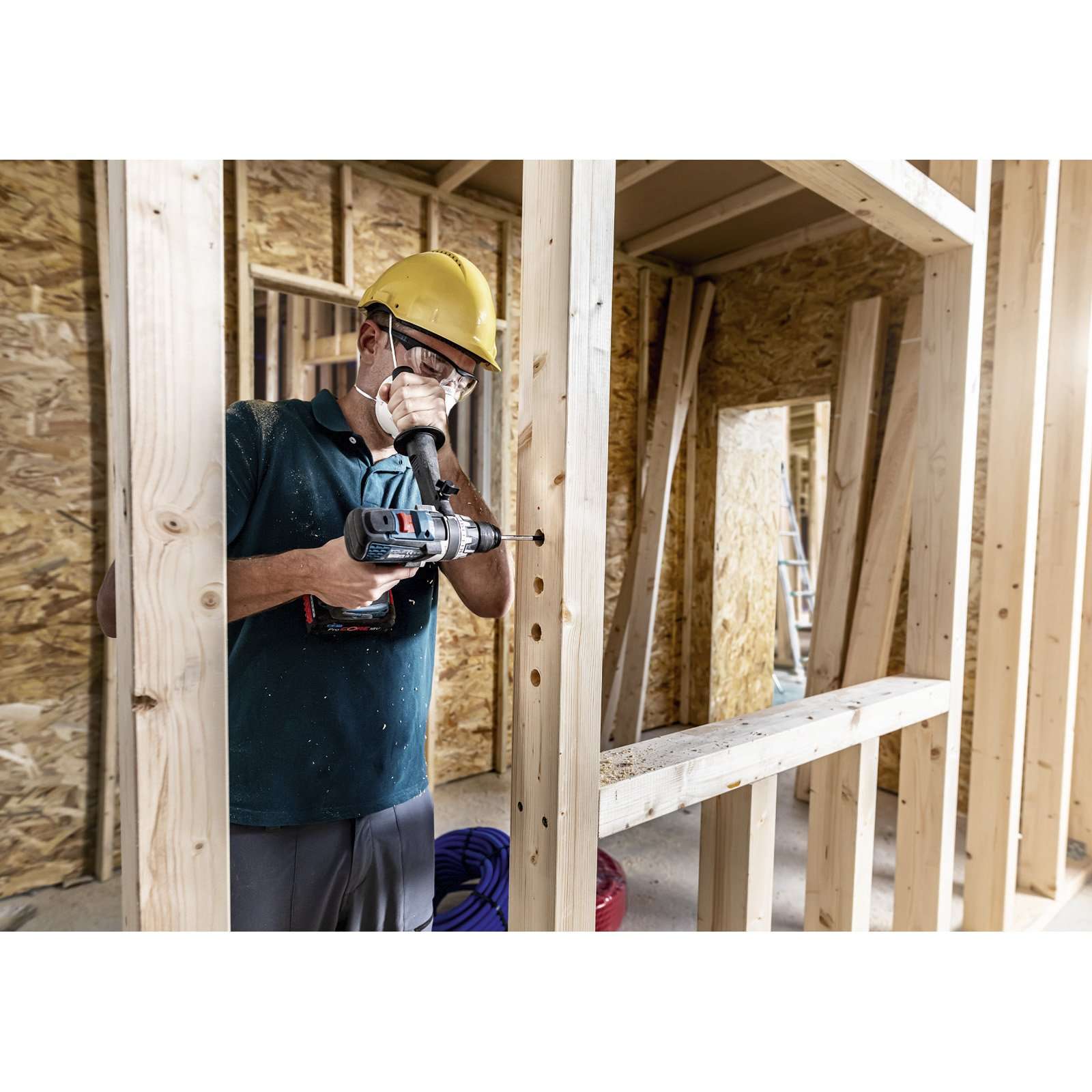 Osoba w żółtym kasku i masce używa wiertarki do zrobienia otworu w drewnianej belce.