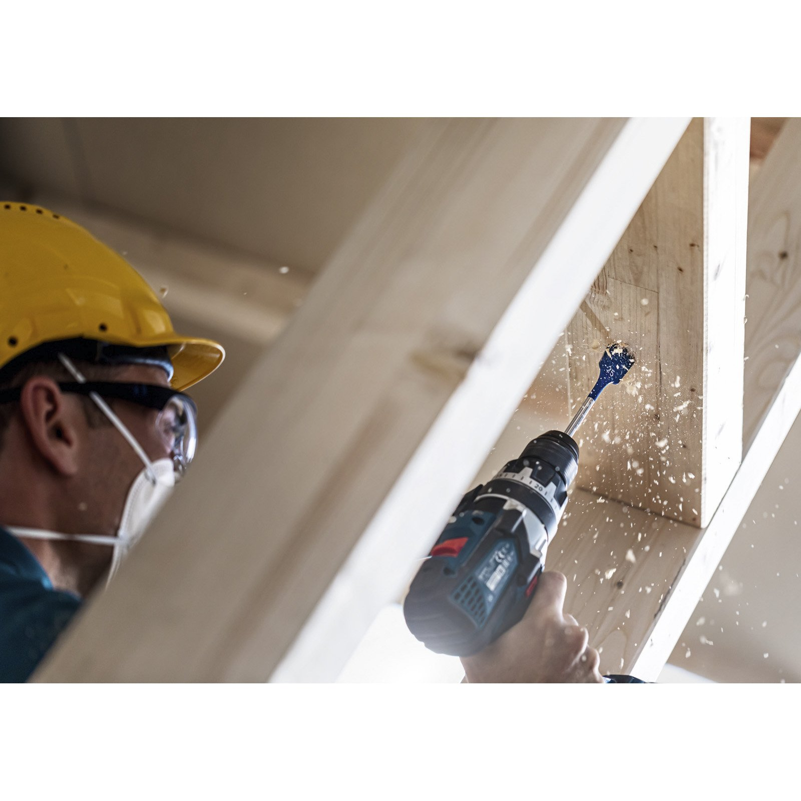 Osoba w żółtym kasku i masce wierci w drewnianej belce. Wióry drewniane wylatują.